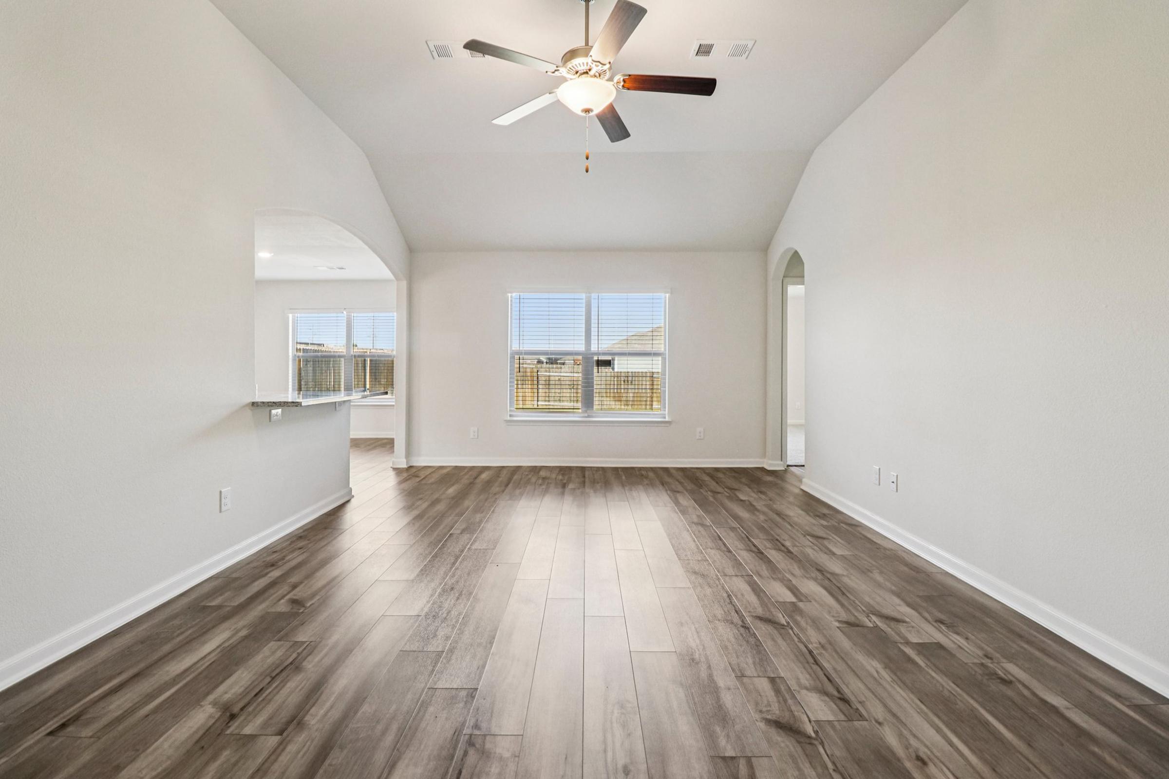 Spacious interior of a modern living room with a ceiling fan, large windows letting in natural light, and wood-look flooring. An adjoining open kitchen area is visible, featuring a bar counter. The walls are painted in a light color, contributing to the bright and airy feel of the space.