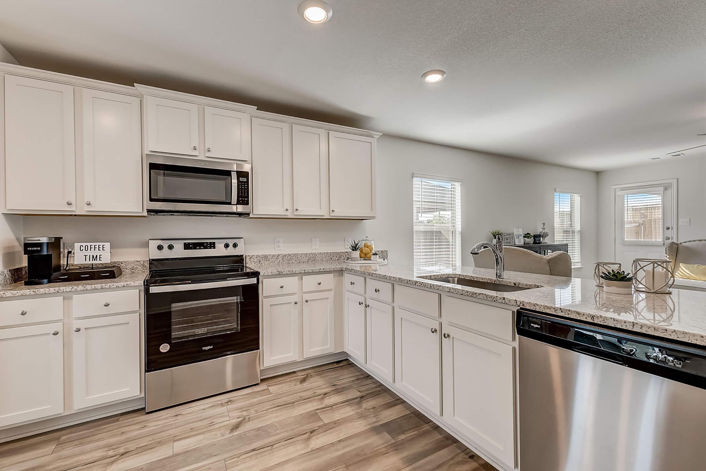Modern kitchen with white cabinetry, stainless steel appliances, and a granite countertop. Features a built-in microwave, oven, and dishwasher. Bright and airy atmosphere, enhanced by natural light from windows. Decorative elements include a "COFFEE TIME" sign and potted plants on the counter.