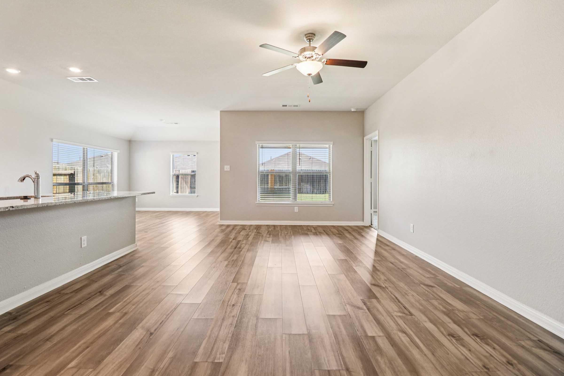 Spacious interior of a modern living room featuring light wood flooring, a ceiling fan, and large windows providing natural light. The open layout showcases a kitchen area with a countertop, creating a bright and inviting atmosphere with access to a backyard view.