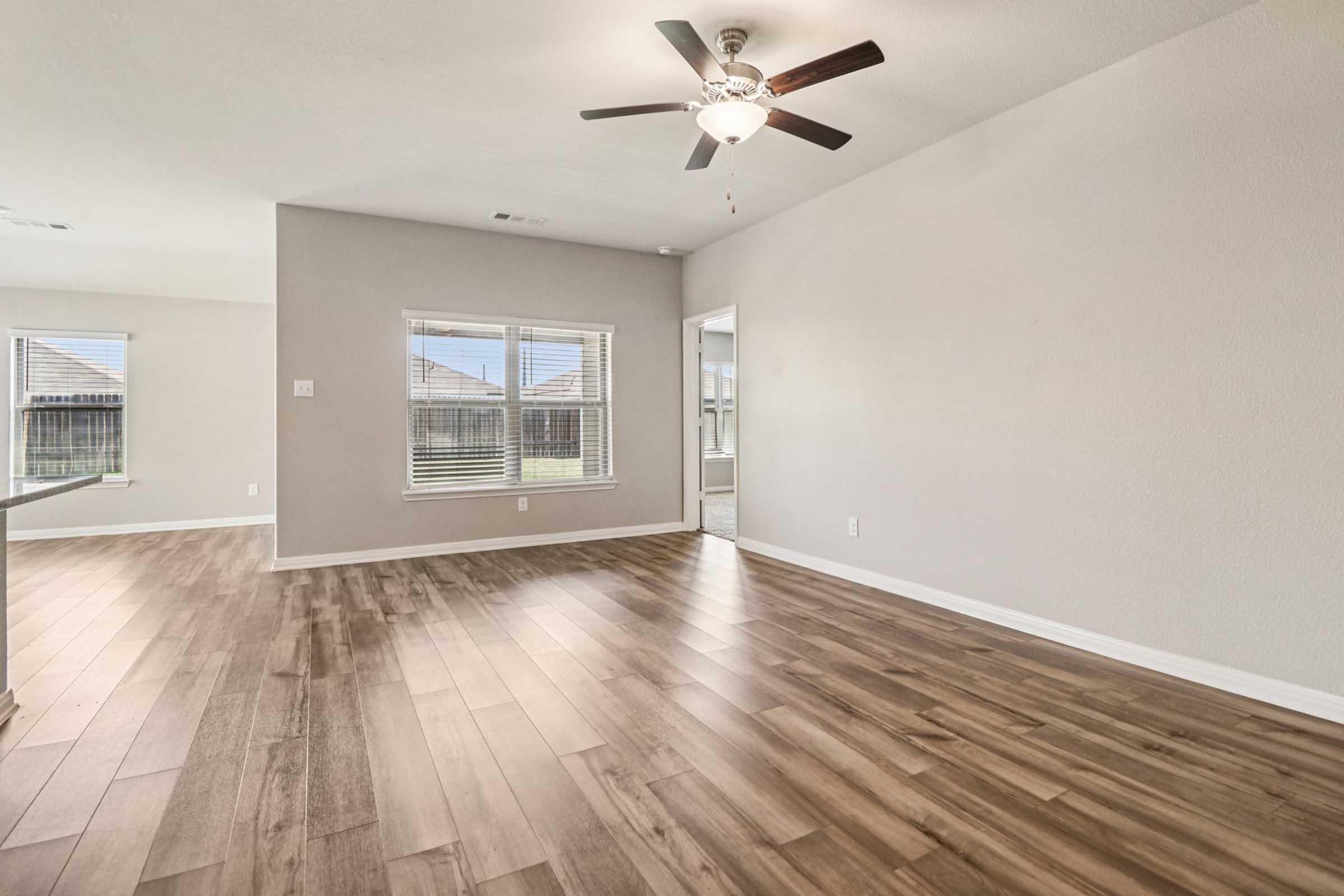 A spacious, empty living room featuring light-colored walls and a wood-paneled floor. A ceiling fan is installed, and natural light enters through multiple windows. The layout is open, leading to a doorway on one side, with no furniture present, creating a clean and airy atmosphere.