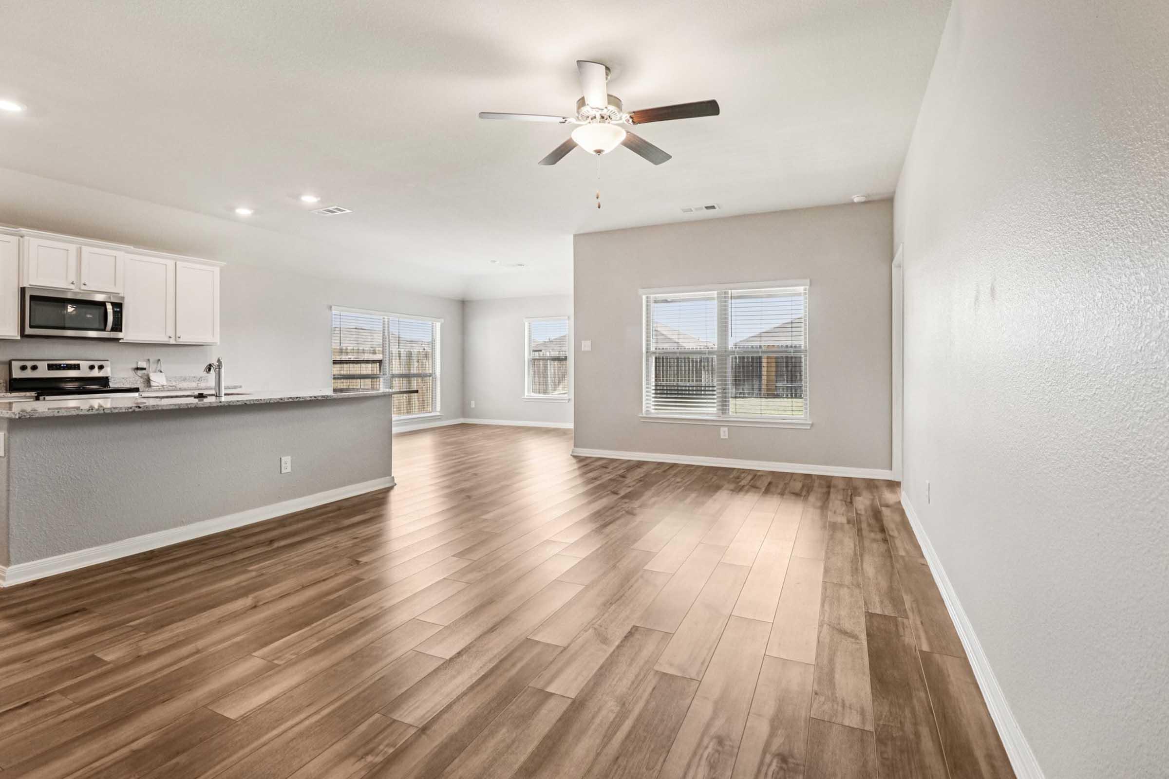 A spacious living area featuring wooden flooring, light gray walls, and a ceiling fan. Large windows allow natural light to fill the room. To the left, there is a modern kitchen with white cabinetry and stainless steel appliances. The space is open and inviting, suitable for various furniture arrangements.