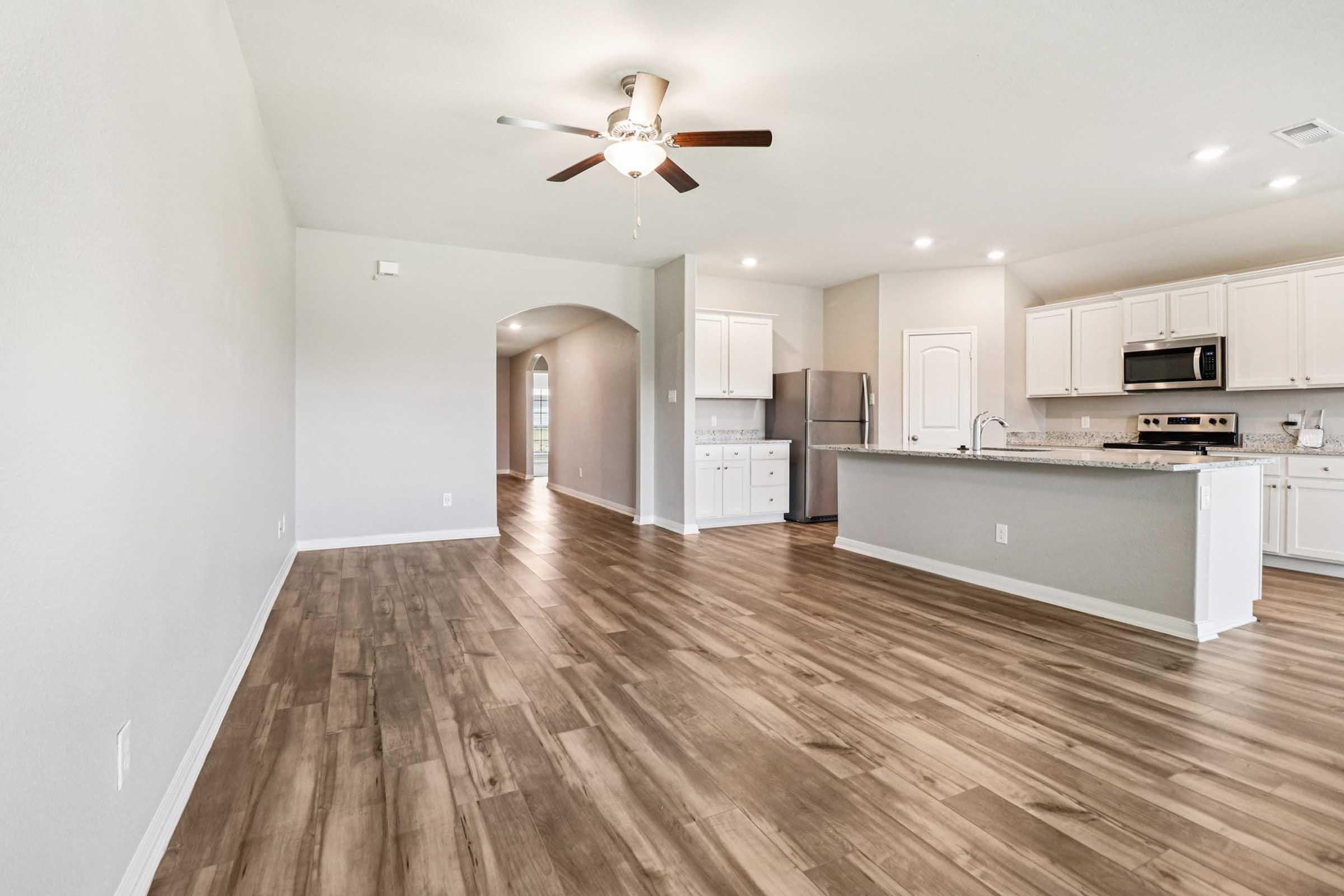 Spacious interior of a modern home featuring a large living area with light wood flooring, a ceiling fan, and an open kitchen with white cabinets and stainless steel appliances. Bright and airy atmosphere with neutral-colored walls and natural light coming from the windows.
