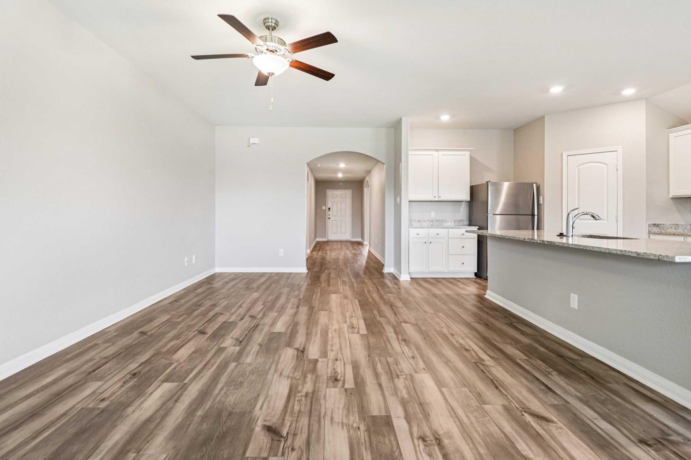 Spacious interior of a modern home featuring a living area with wooden flooring, a ceiling fan, and an open layout. The kitchen is visible in the background, equipped with white cabinets and stainless steel appliances. Natural light brightens the space, enhancing the inviting atmosphere.