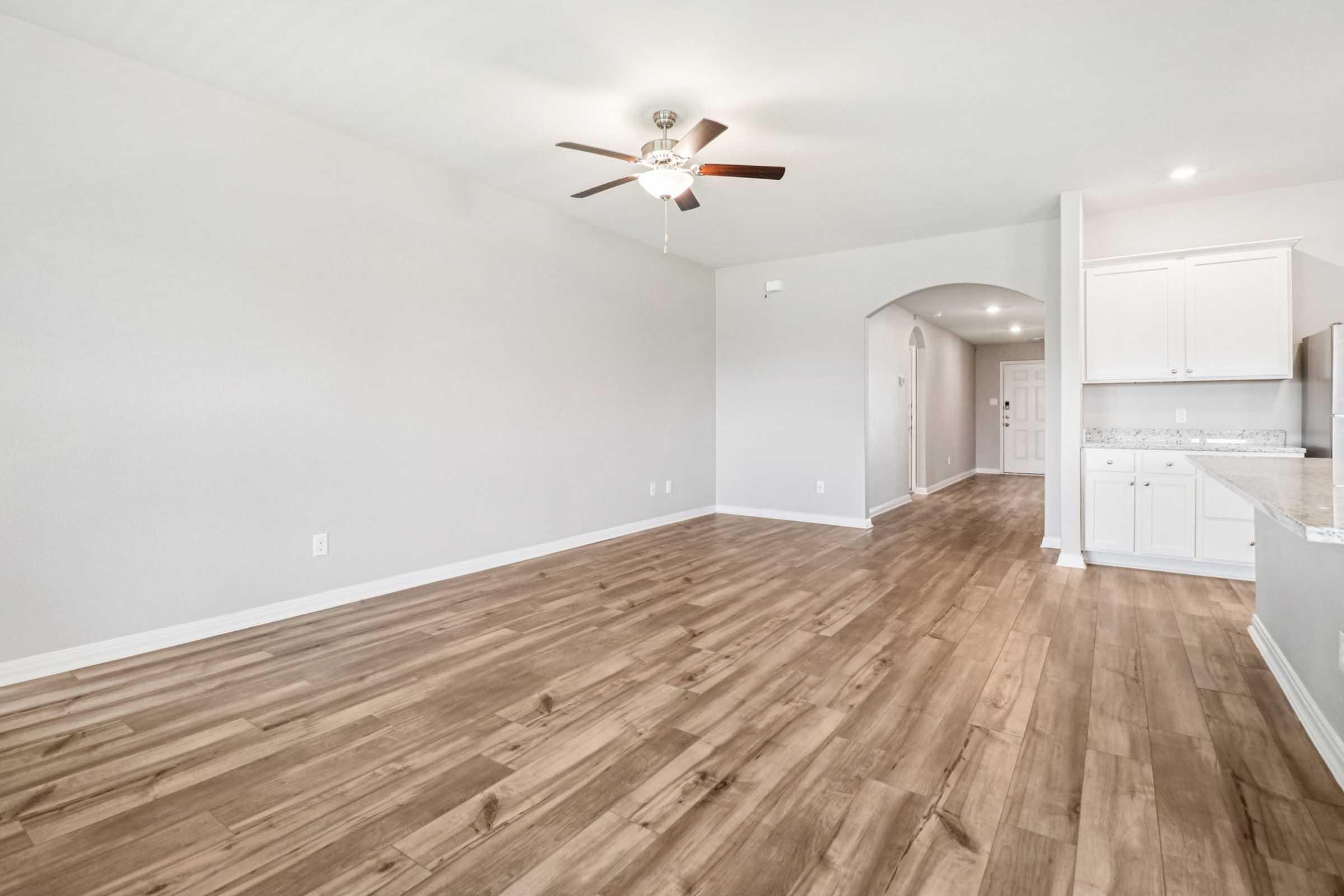 Interior view of a spacious, well-lit room featuring light-colored walls, a ceiling fan, and hardwood-style flooring. The area includes a kitchen to the right with white cabinetry. An archway leads to a hallway, creating an open and inviting atmosphere.