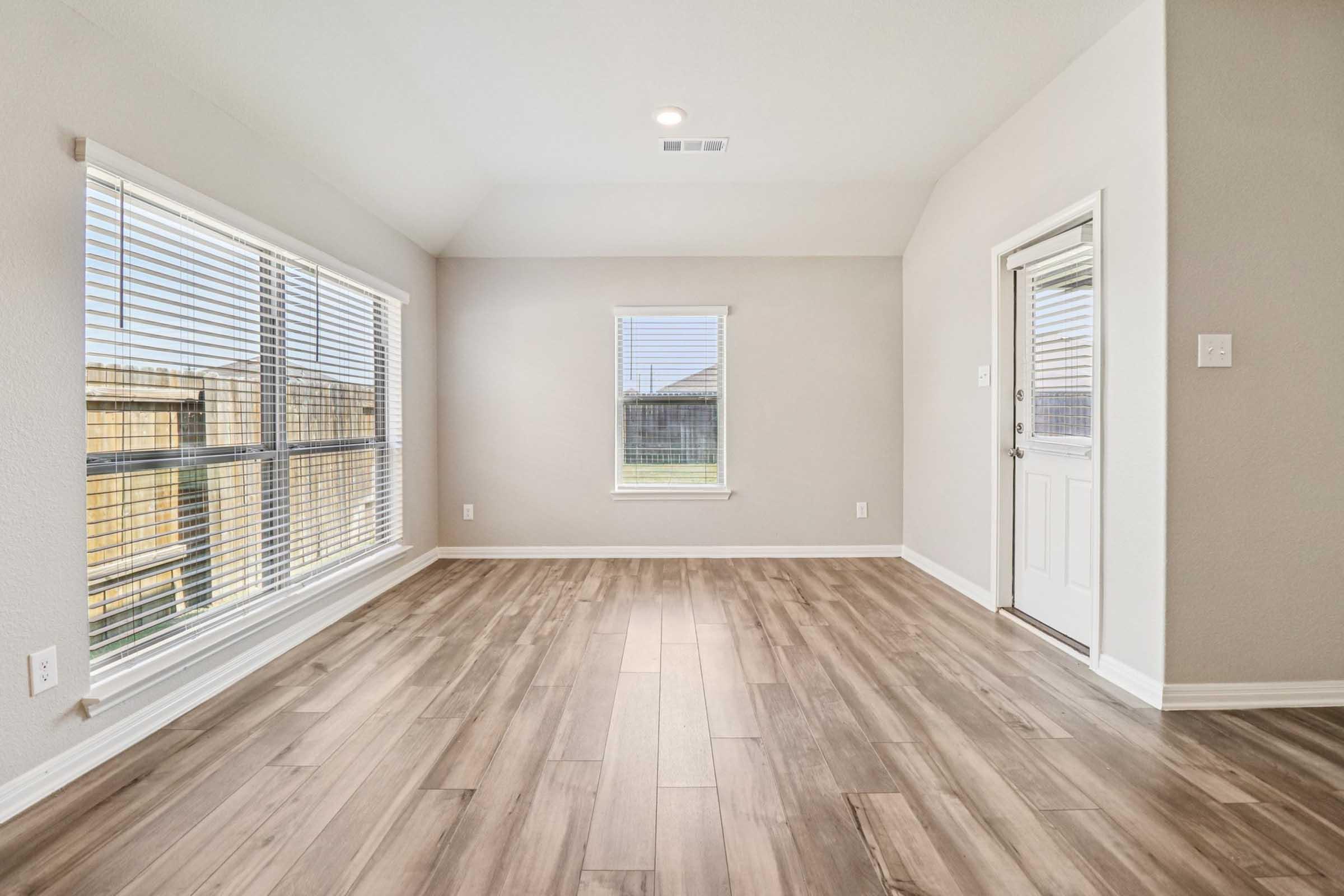 A bright, empty room with large windows featuring blinds and light-colored walls. The floor is made of wood-like tiles, and there is a door leading outside. A window without blinds is also visible, allowing natural light to enter. The room has a spacious and clean appearance.