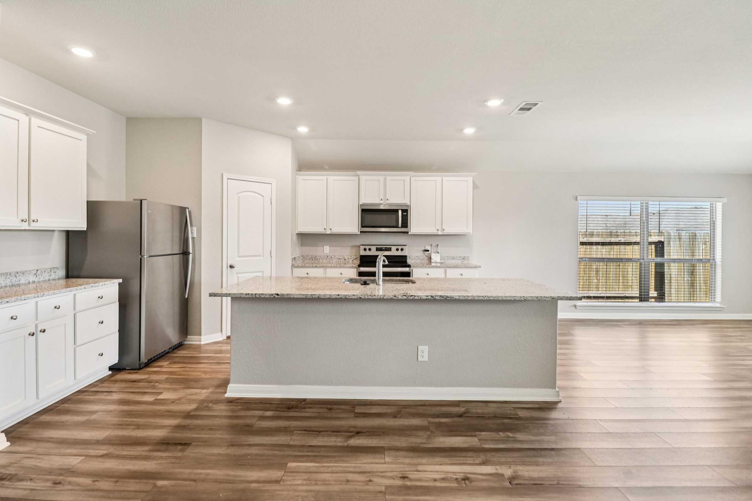 Modern kitchen featuring white cabinets, granite countertops, stainless steel appliances, and an island with a sink. The open layout includes a large window allowing natural light, and wooden flooring throughout the space. Ideal for cooking and entertaining.