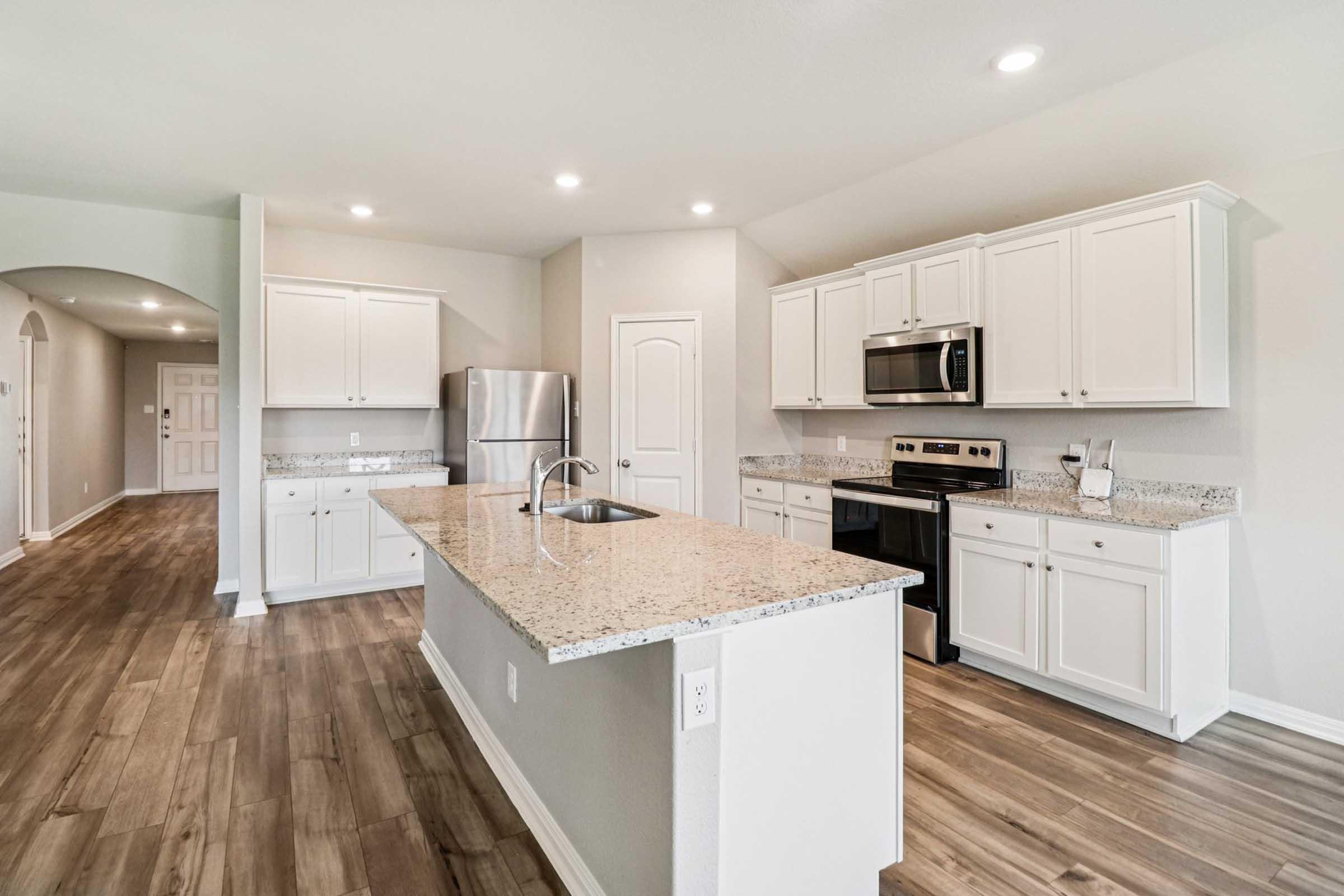 Bright and modern kitchen featuring white cabinetry, granite countertops, stainless steel appliances, and a large island. The open layout includes a spacious dining area, light hardwood flooring, and soft recessed lighting, creating an inviting atmosphere. A hallway leads to the entrance.
