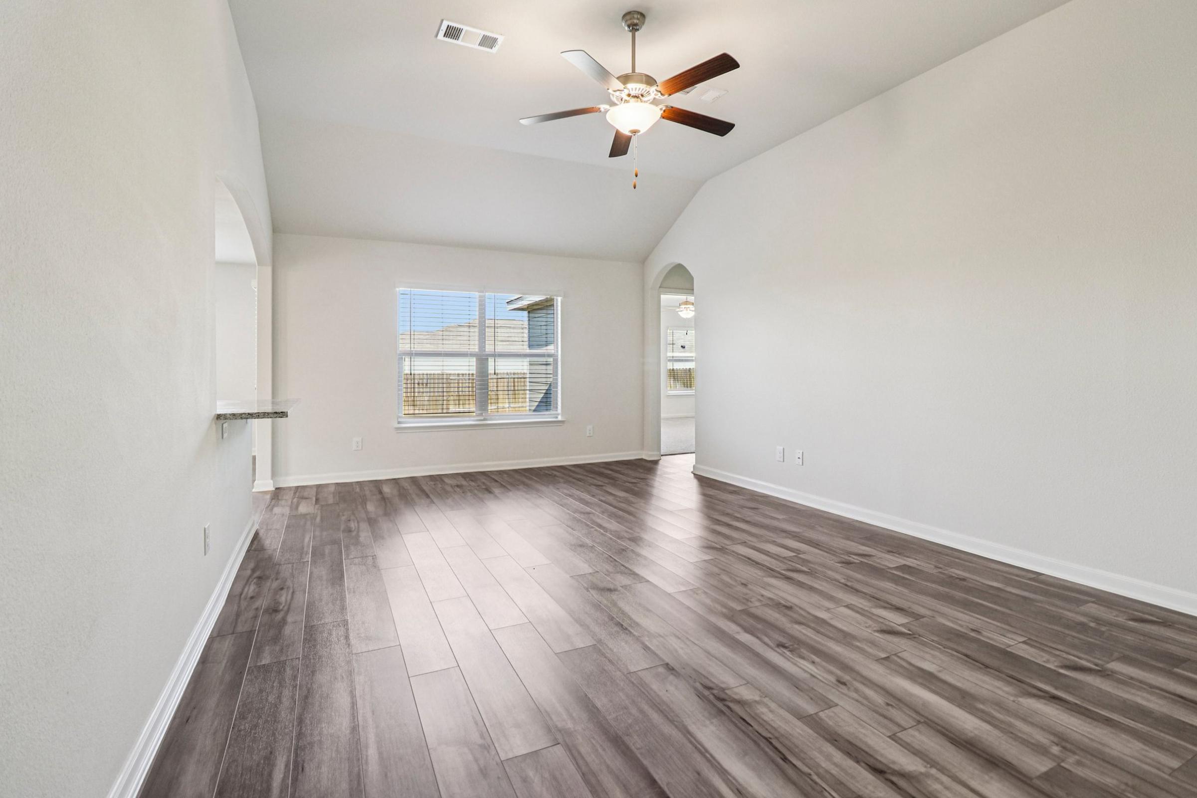 Spacious interior of a modern home featuring light-colored walls, a ceiling fan, and large windows allowing natural light. The floor is covered with dark wood-like tiles, and there is an arched doorway leading to another room. The overall atmosphere conveys a clean and inviting space.