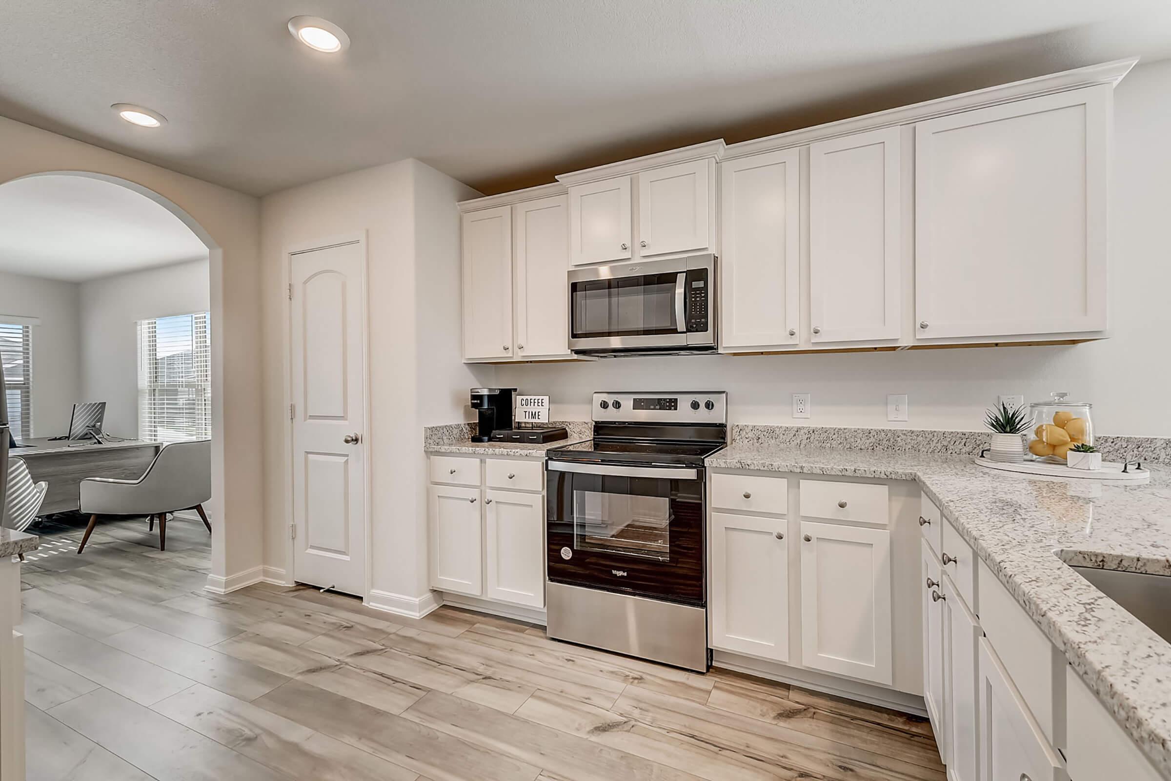 Modern kitchen with white cabinetry, stainless steel appliances, granite countertops, and a spacious layout. Features include a microwave, oven, coffee maker, and an arched entrance leading to a living area with natural light from large windows. A small decorative plant and fruit bowl add a homey touch.