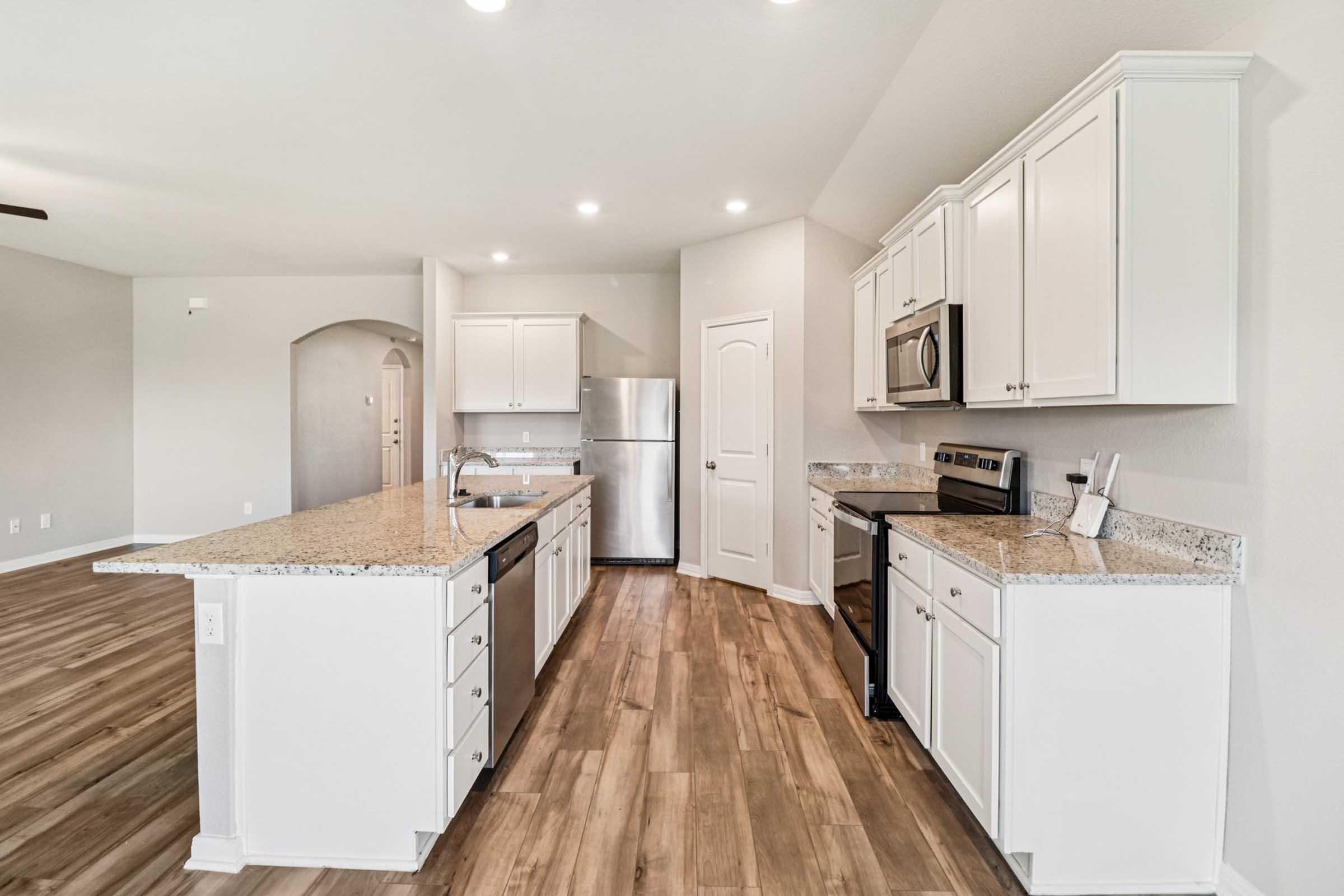 A modern kitchen featuring white cabinetry, granite countertops, stainless steel appliances, and a spacious layout. The kitchen includes an island with a sink, a built-in microwave, an oven, and a fridge. Bright overhead lights illuminate the room, which has a neutral color scheme and hardwood flooring.