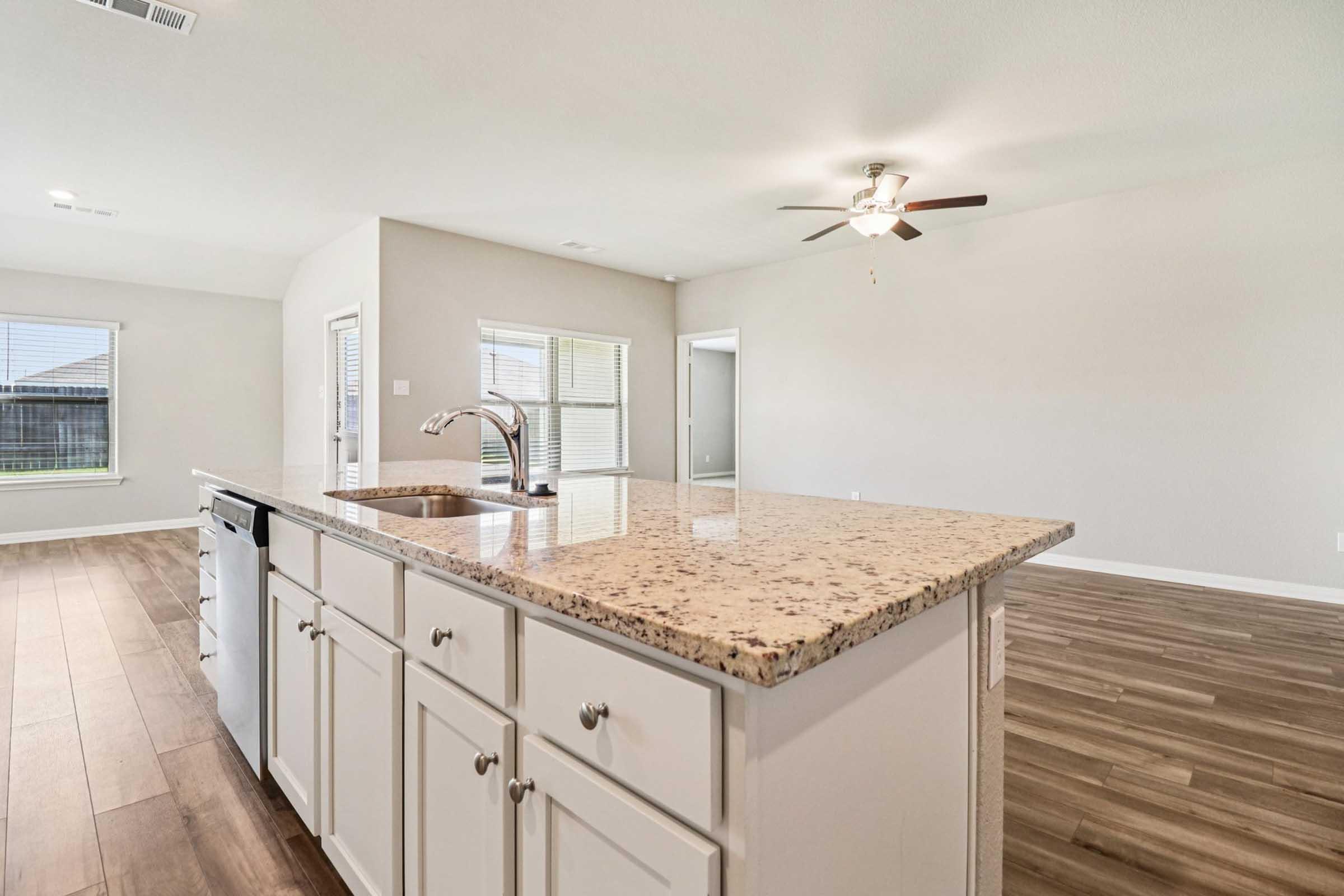 Modern kitchen featuring a spacious island with a granite countertop, white cabinetry, and stainless steel appliances. The open layout showcases a living area with wood-style flooring and a ceiling fan, with windows providing natural light and a view of the outside.