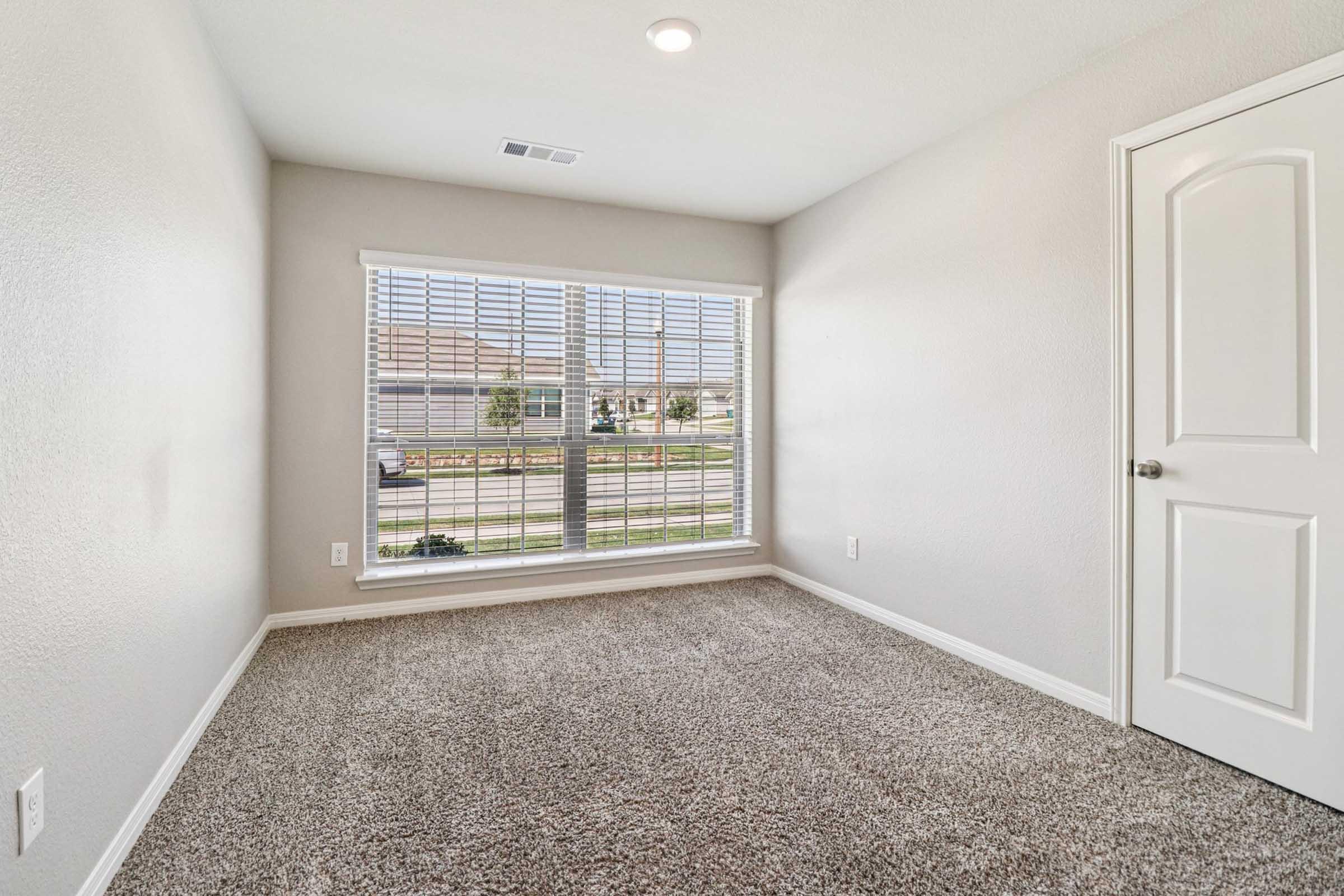 An empty room with light gray walls and carpeted flooring. A large window with horizontal blinds allows natural light inside, revealing a view of a street outside. A closed white door is visible on the right side of the image. The room has a clean and minimalist appearance.