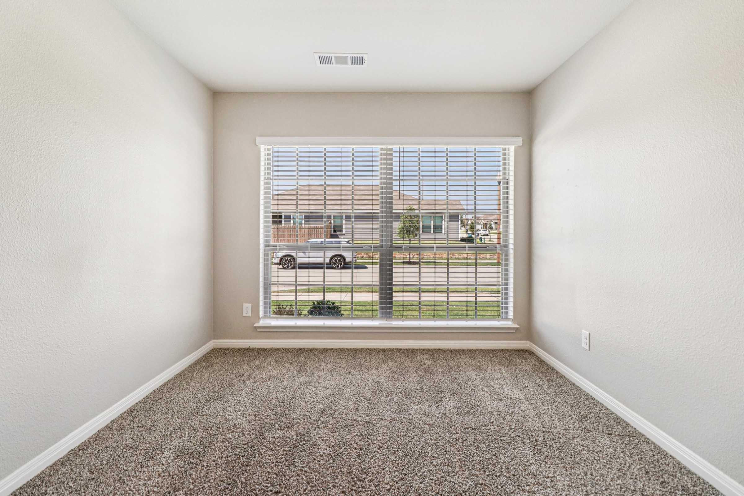 A minimalist empty room with light beige walls and carpet. A large window with white blinds allows natural light in, showcasing a view of a grassy area and a street outside. No furniture is present, emphasizing the spaciousness of the area.