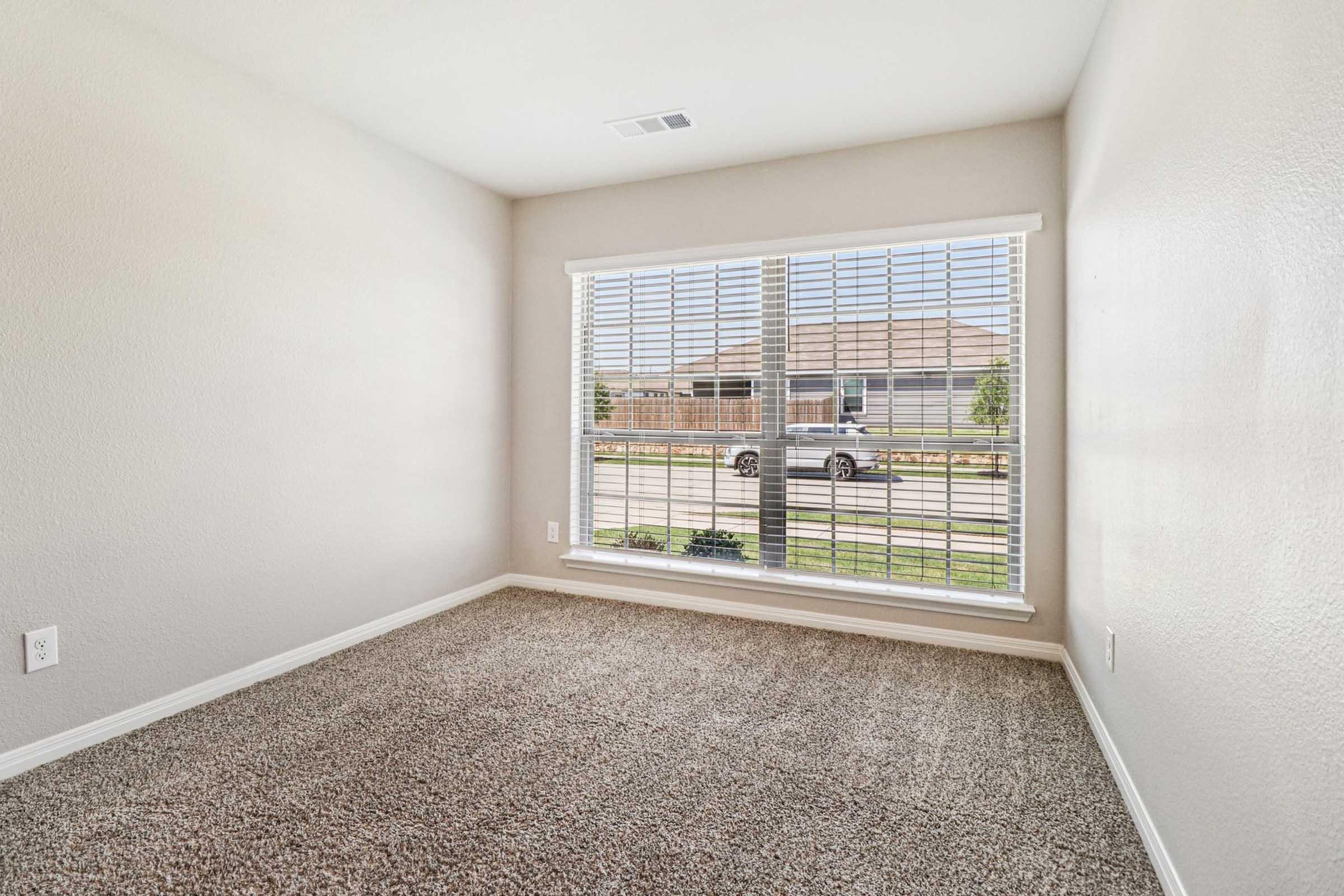 A bright, empty room with beige walls and carpet flooring. A large window with white blinds allows natural light to enter, providing a view of the outside. The space is uncluttered, emphasizing its potential for various uses, such as a bedroom or office.