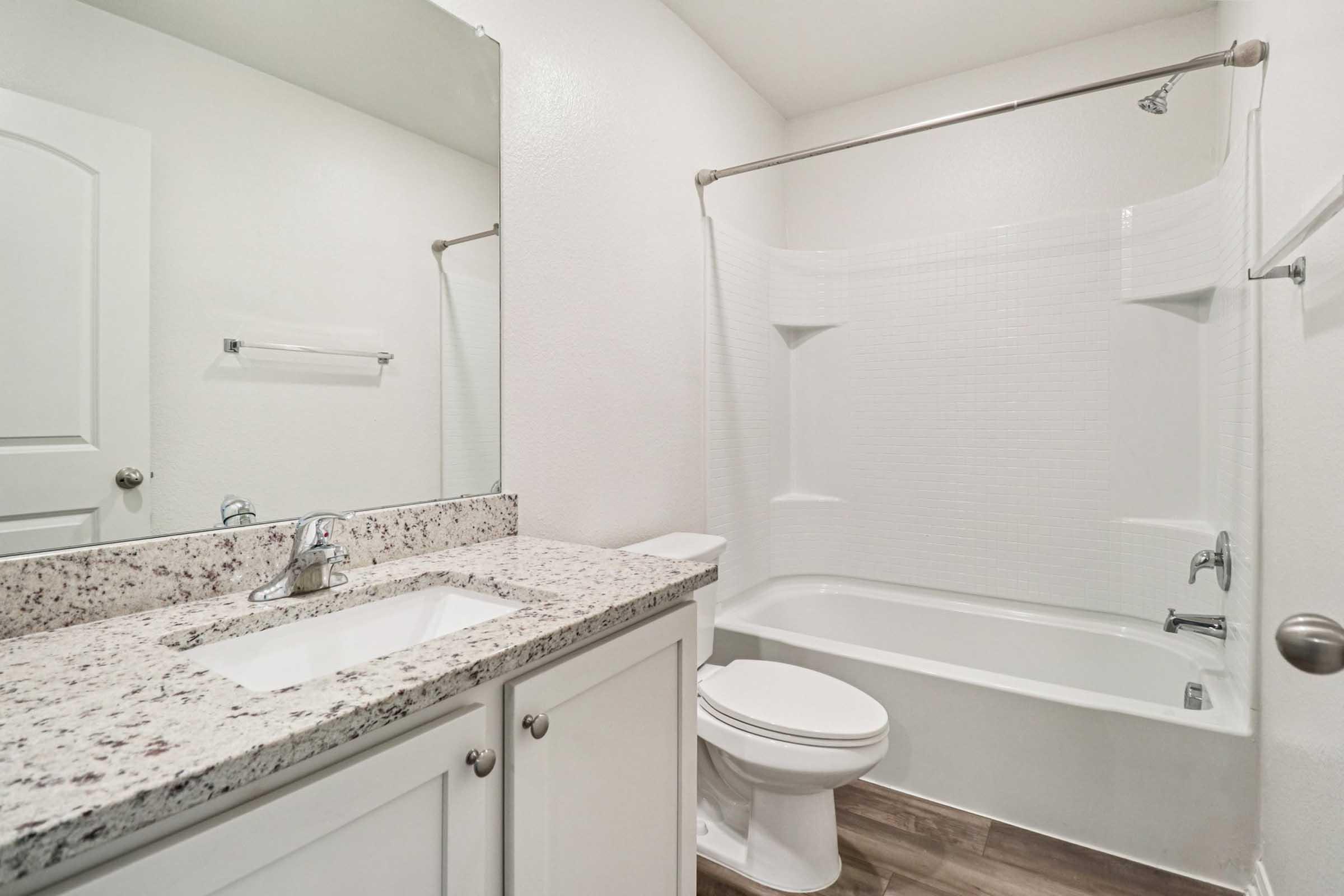 A clean, modern bathroom featuring a granite countertop sink, a mirror, and a white bathtub with a shower curtain. The walls are painted in a light color, and the flooring is a wood-like tile. A towel rack is visible, and a door to the left leads to another room.