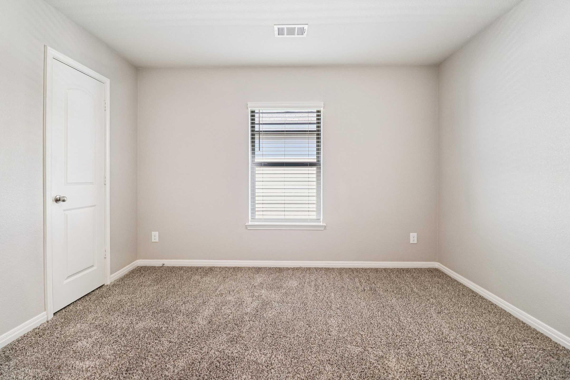 A light-colored vacant room featuring beige walls and plush carpet. There's a single window with blinds on the left and a closed door on the right. The space is well-lit, highlighting its emptiness and simplicity, making it suitable for various uses.