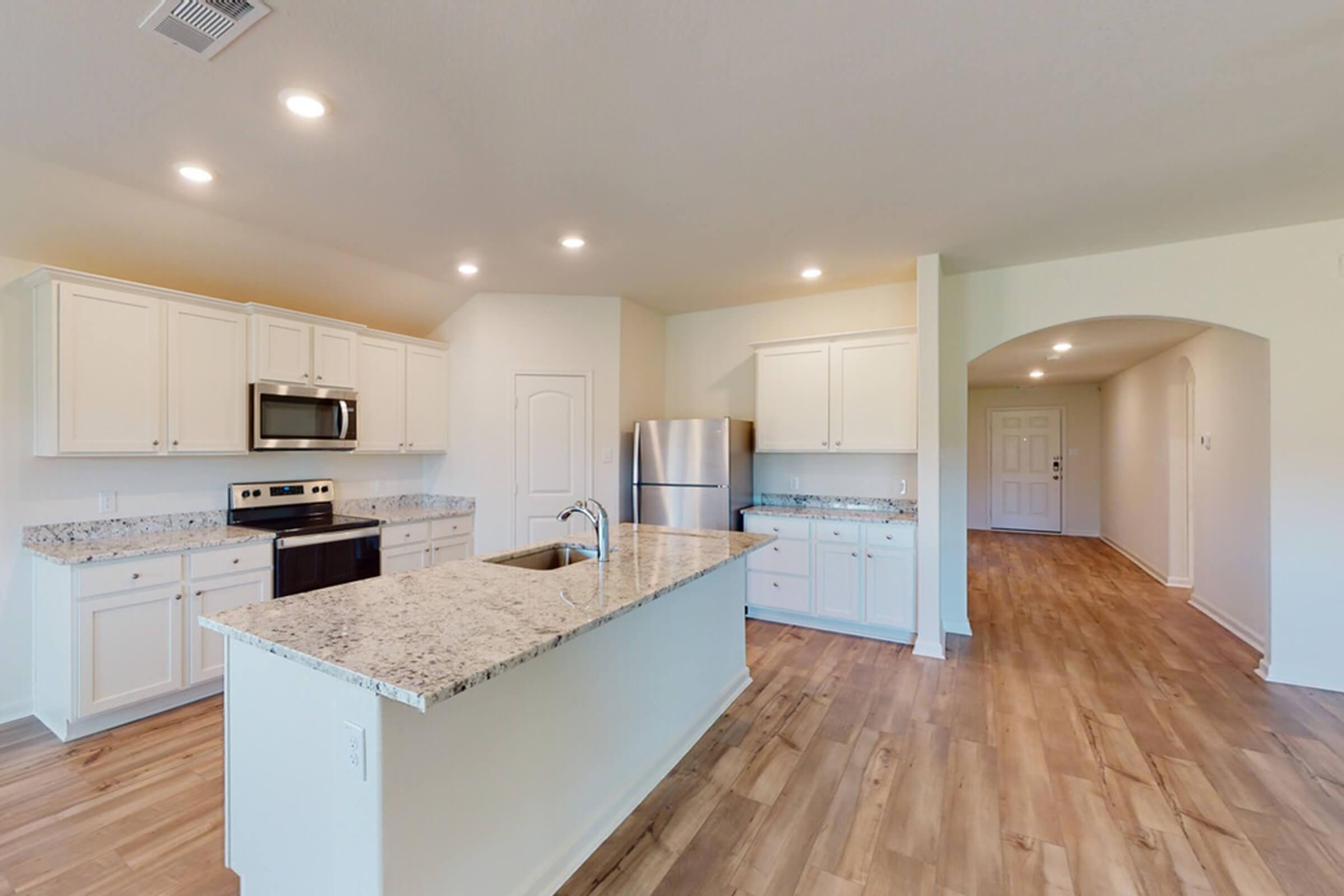 A modern kitchen with white cabinetry and granite countertops. It features stainless steel appliances, including a microwave and refrigerator. An island with a sink is at the center, and open space leads to a hallway with a front door. The floor is covered in light wood planks, creating a bright and airy atmosphere.