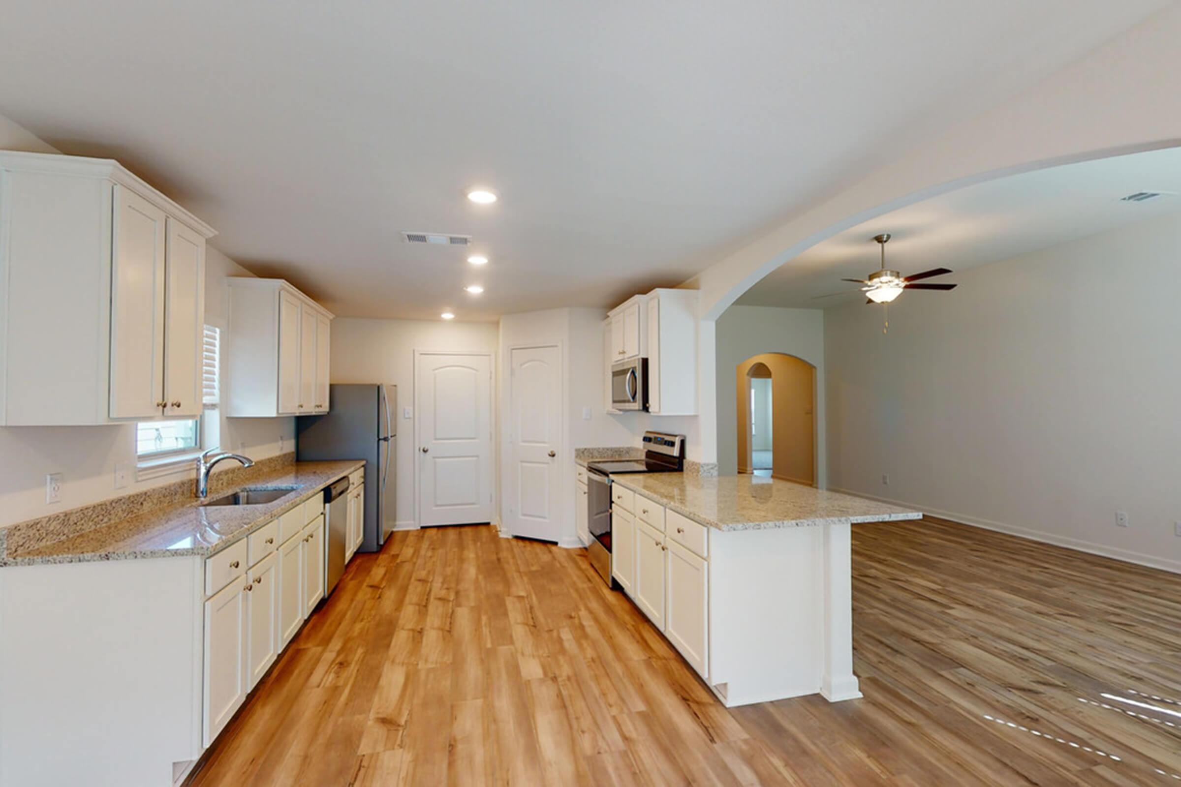 Modern kitchen with light-colored cabinetry and granite countertops, featuring stainless steel appliances. A spacious layout connects to a living area with wooden flooring and a ceiling fan. Natural light enters through windows, creating a bright atmosphere.