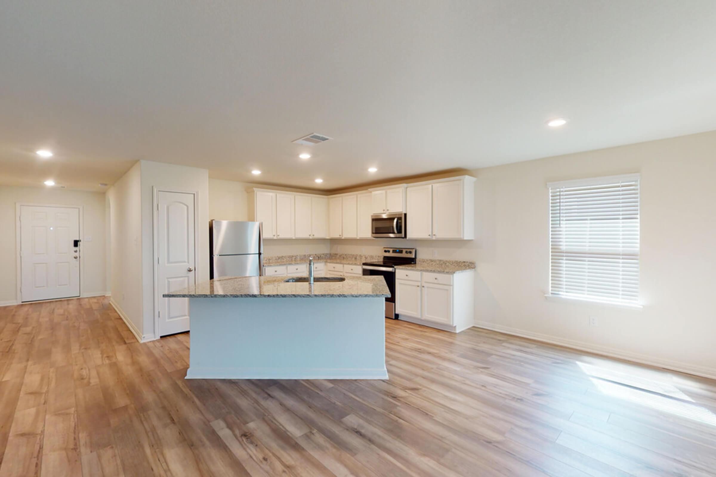 Modern kitchen featuring white cabinetry, stainless steel appliances, and a spacious island with a granite countertop. The room has hardwood flooring and large windows that provide natural light. A door on the left leads to an entryway.