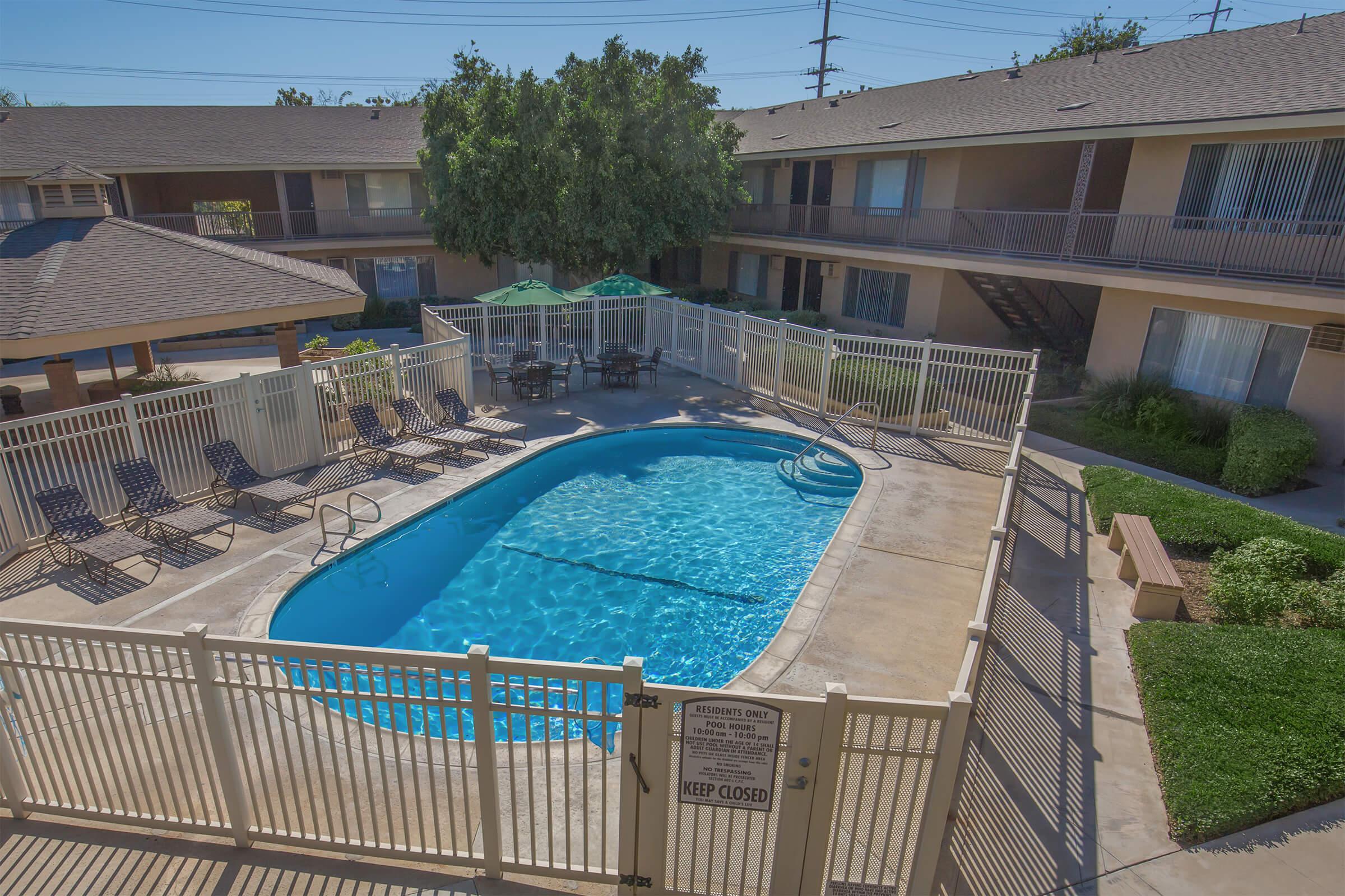 Aerial view of a swimming pool surrounded by a white fence in an outdoor space with lounge chairs. Nearby are green umbrellas and tables for relaxation. The area includes two-story apartment buildings with balconies, with trees and bushes adding greenery to the environment. A sign states "KEEP CLOSED" near the pool area.