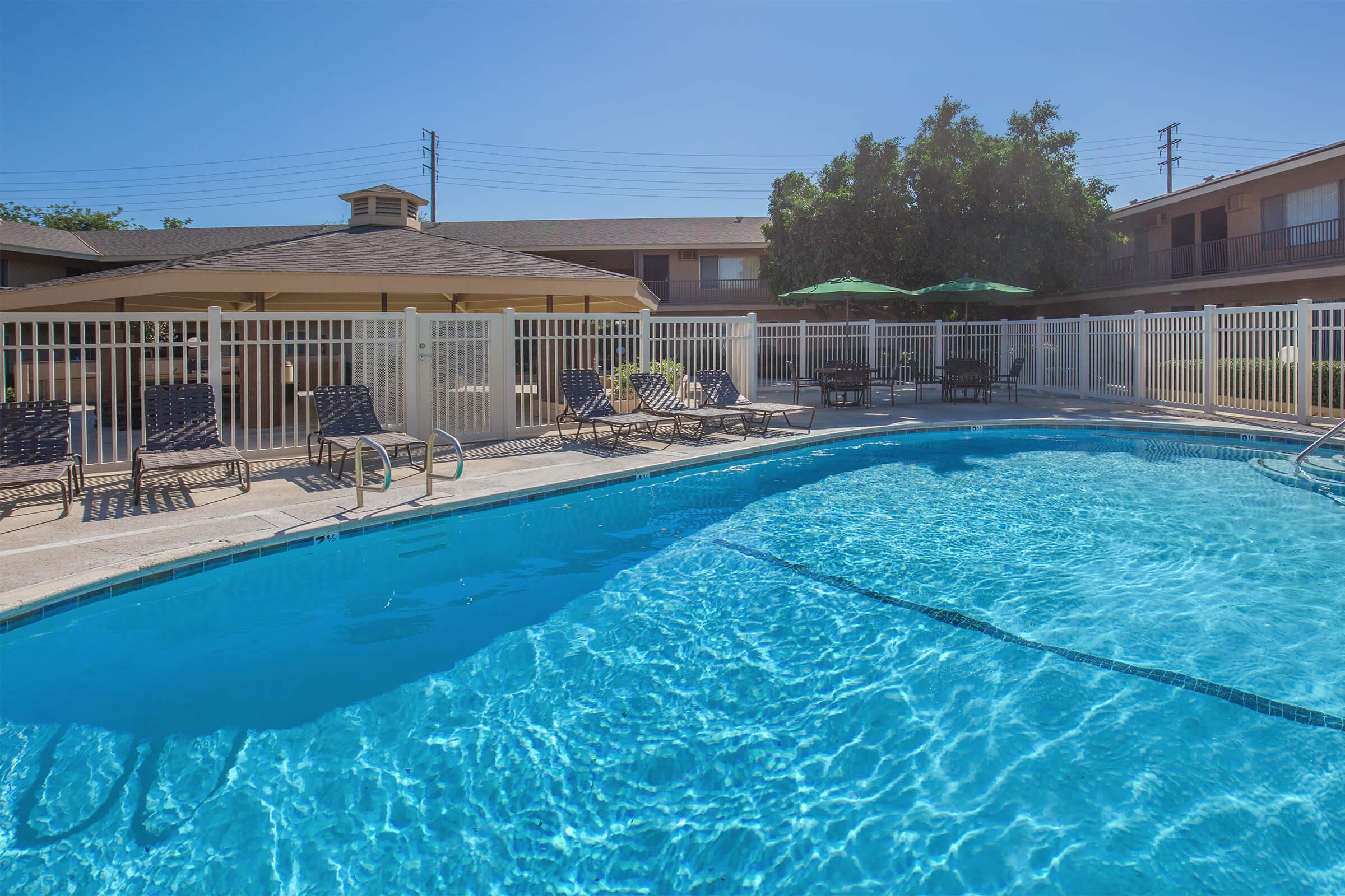 Swimming pool surrounded by a fenced area with lounge chairs and umbrellas. In the background, there are buildings and a gazebo with additional seating. The sky is clear and blue, creating a sunny, inviting atmosphere.