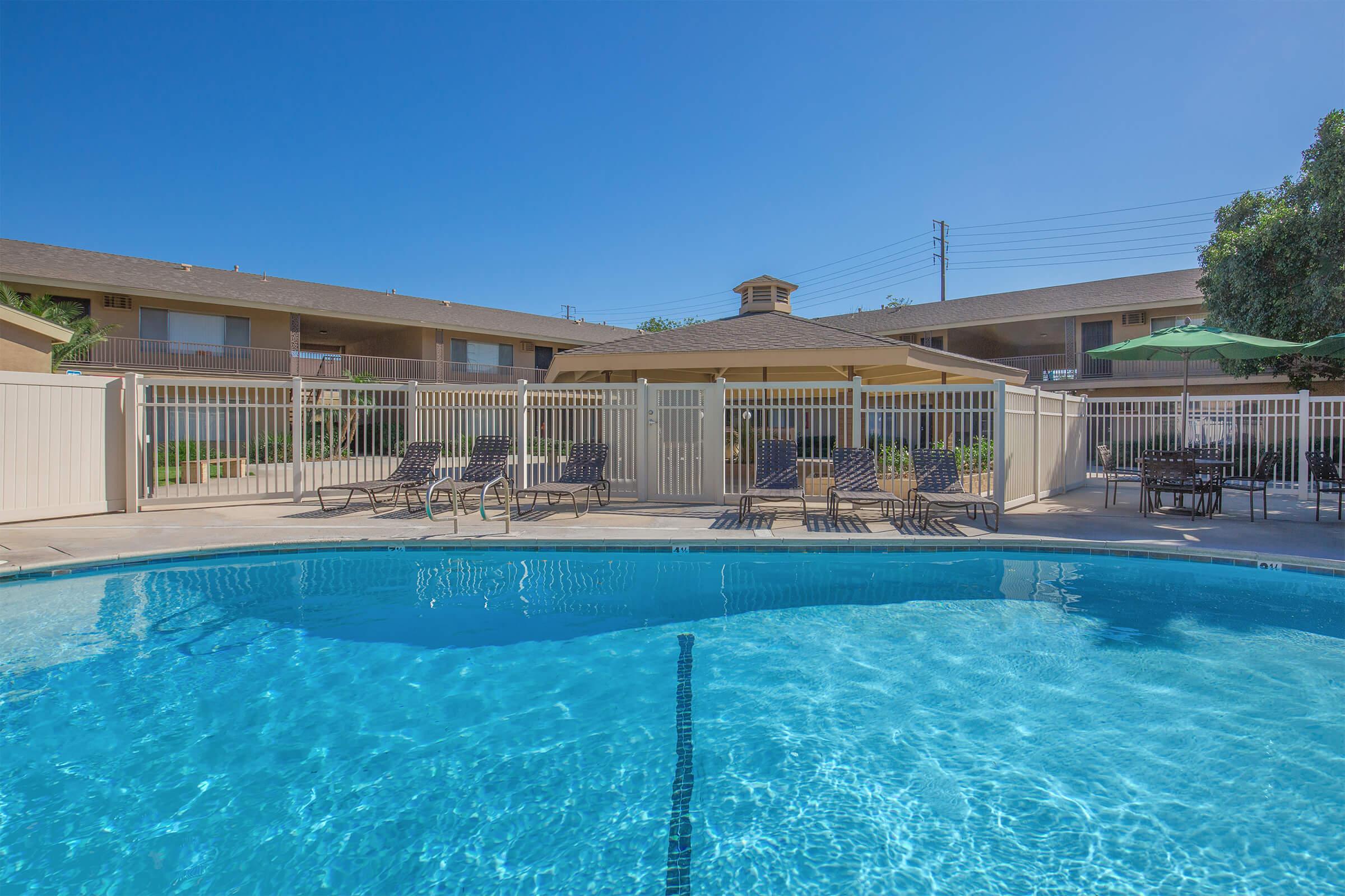 A clear blue swimming pool surrounded by lounge chairs and a fence. In the background, there are two-story buildings with balconies, and a shaded area with an umbrella. The sky is bright and cloudless, creating a sunny, inviting atmosphere.