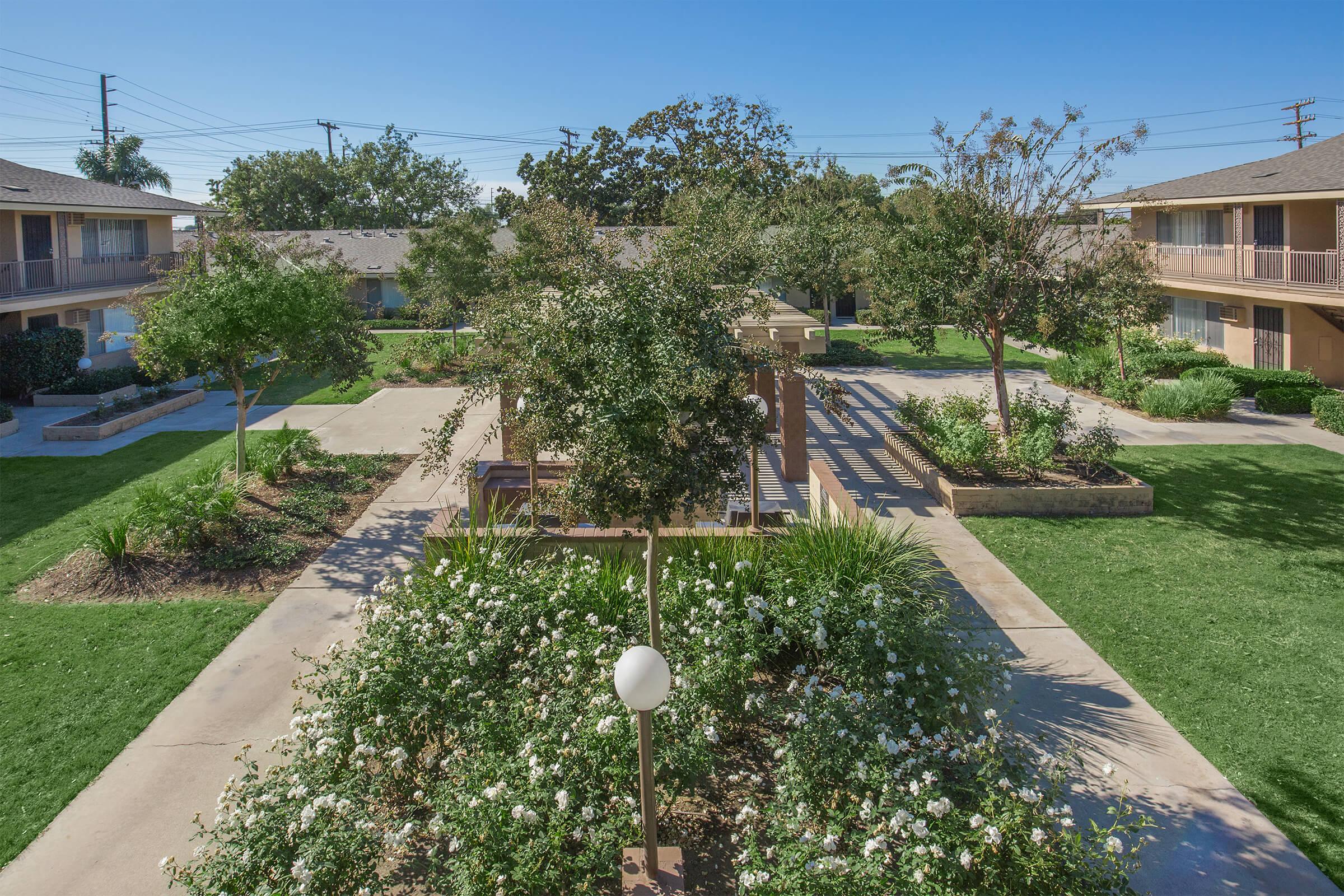 A landscaped courtyard featuring lush greenery, flower beds, and a central pergola, surrounded by low-rise apartment buildings. The scene is well-lit by sunlight, with trees providing shade and a clear blue sky above.