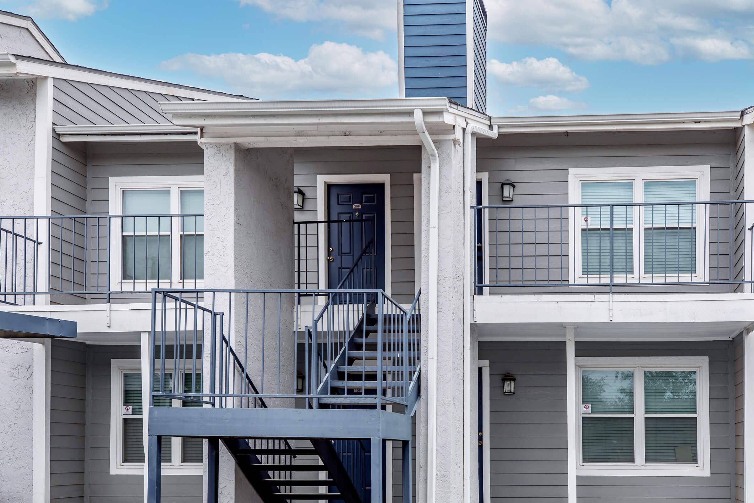 Two-story apartment building with gray siding. Features a central blue door and white window trim. A metal staircase leads to the second floor, with balconies visible on both levels. The building is set against a blue sky with scattered clouds.