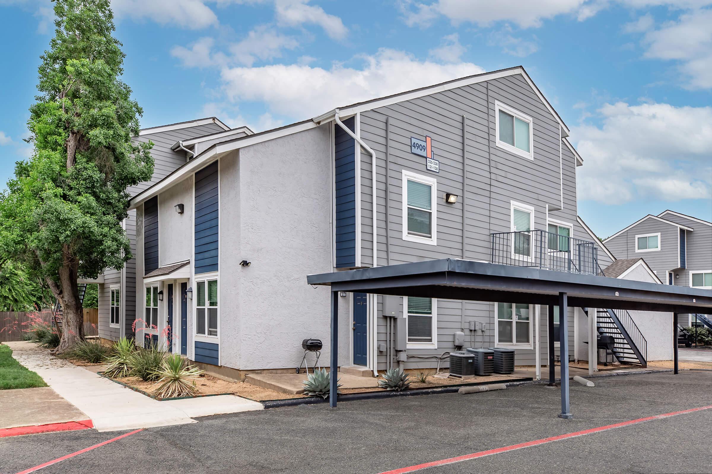 A modern two-story apartment building with a mix of grey siding and white accents. It features a covered parking area, a staircase leading to the upper floor, and surrounding landscaping with small plants. The sky is partly cloudy, adding to the inviting atmosphere of the property.