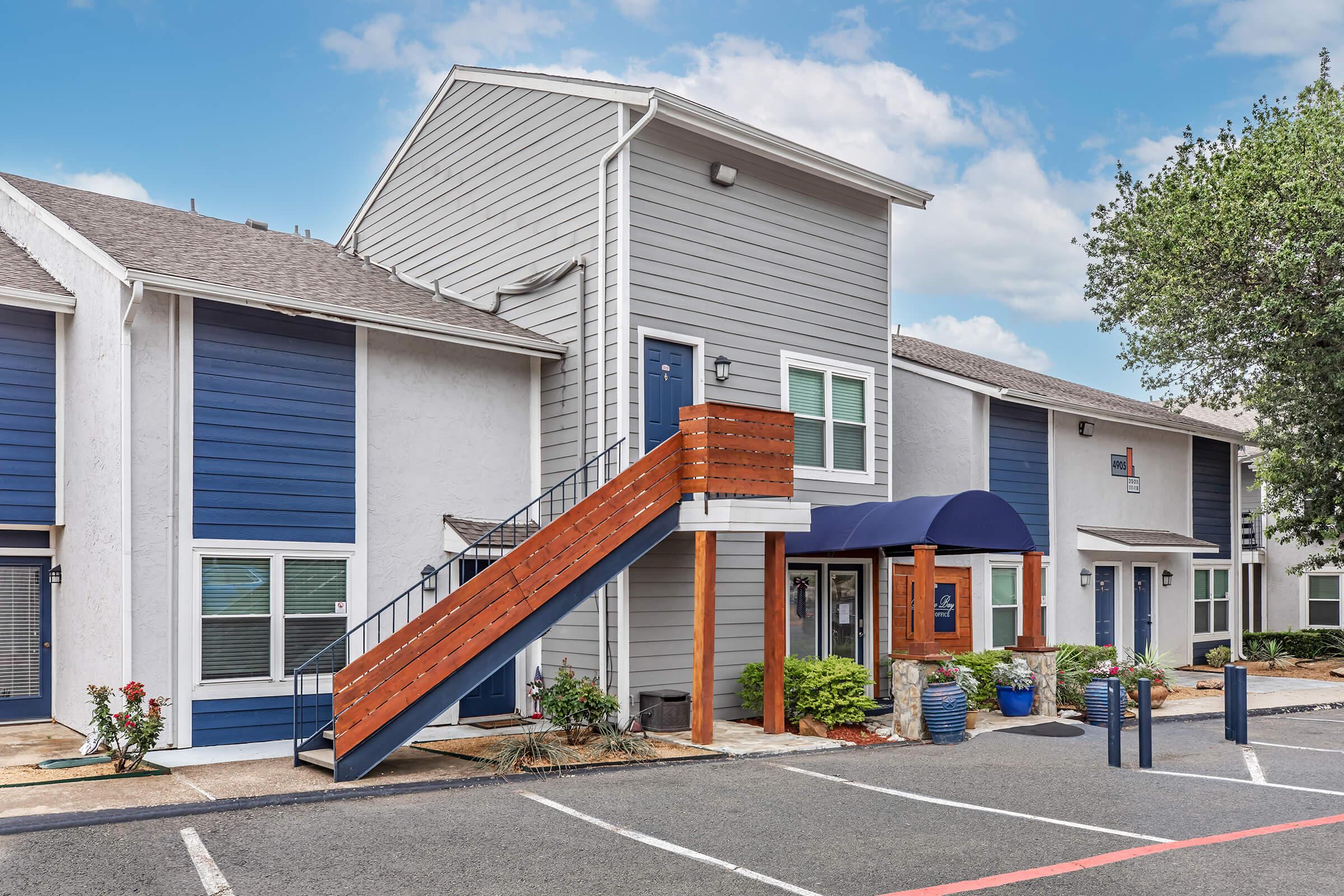 Two-story apartment building featuring a combination of blue and gray siding. A wooden staircase leads to the second floor, and a covered entrance is visible on the ground level. The parking lot has landscaping with bushes and small trees, under a partly cloudy sky.