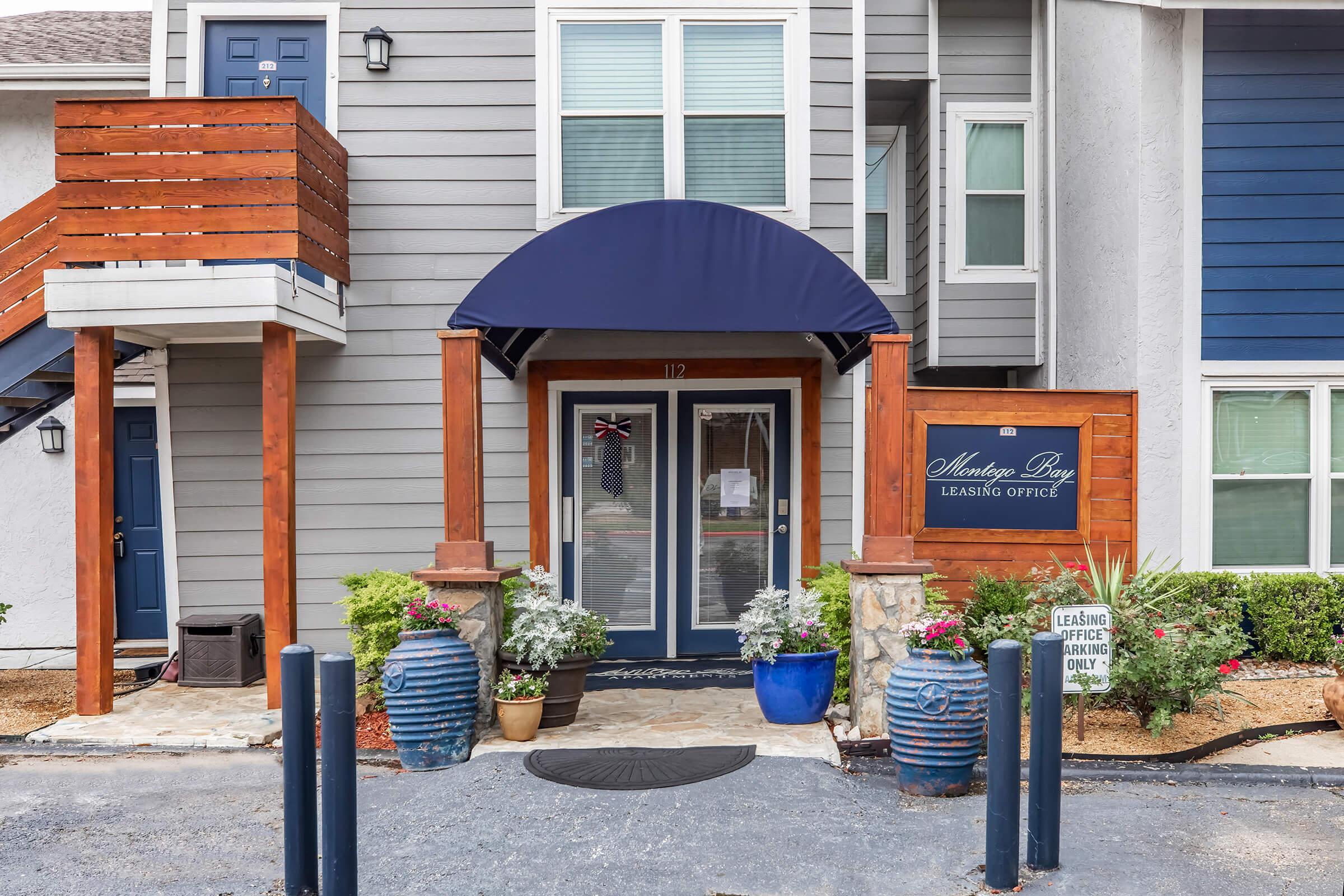 Front view of a leasing office building featuring a blue canopy over the entrance. The entrance is framed by potted plants and decorative features. A sign indicates the leasing office hours, and nearby, there are blue decorative pots. The building has a multi-level design with wooden accents.