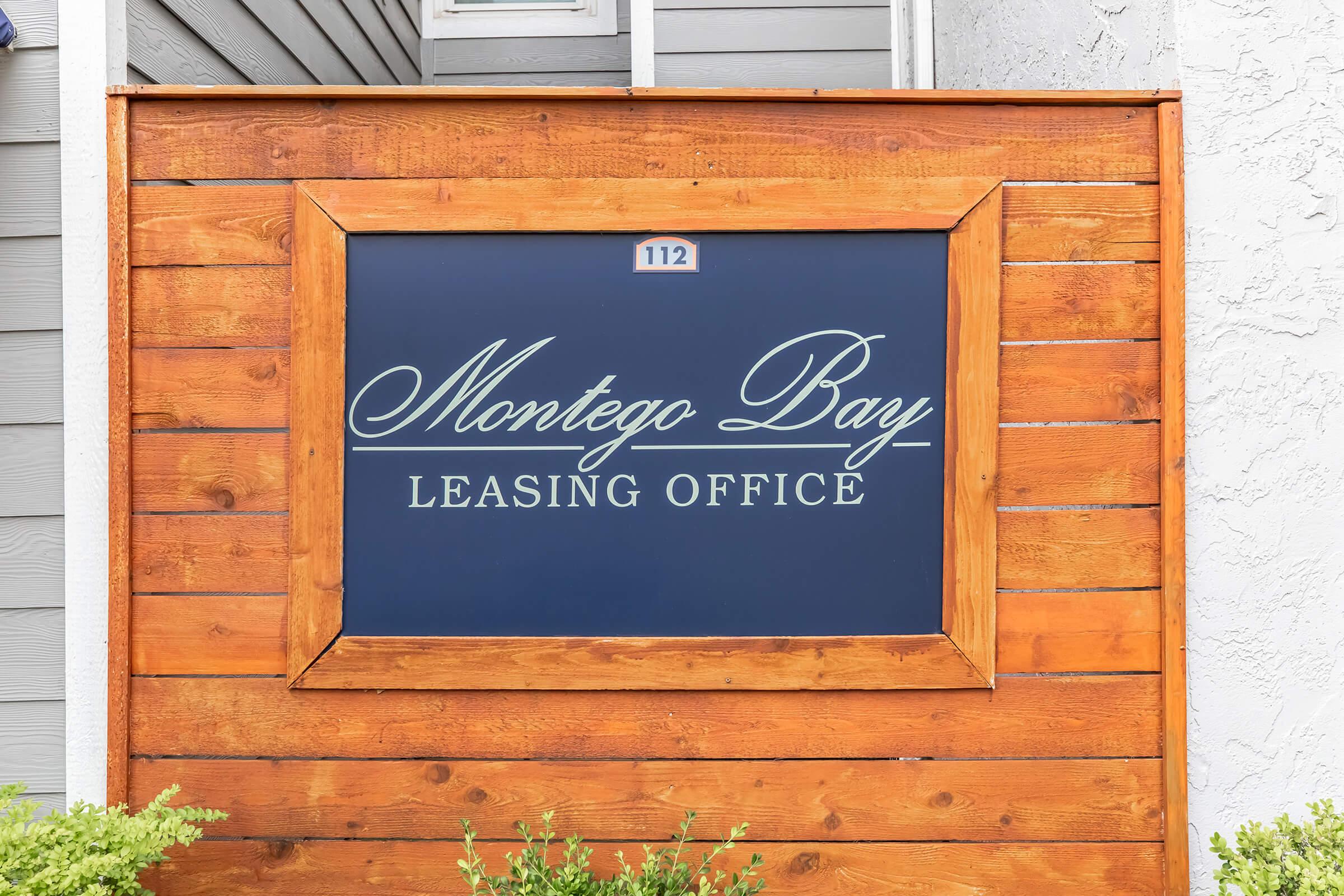 Sign for "Montego Bay Leasing Office" with the number 112, set against a wooden frame. The background is a deep blue with elegant white lettering, surrounded by a wooden structure, conveying a welcoming atmosphere.