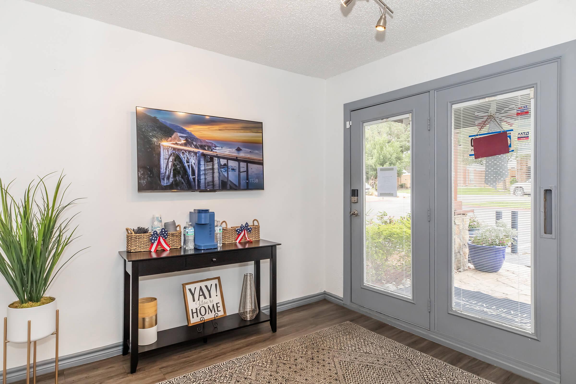 A stylish entryway featuring a console table with a decorative sign saying "YAY! HOME," a plant, and a water dispenser. A large wall-mounted TV displays a scenic landscape, while a bright entrance door with a window offers a view of the outside. The floor is adorned with a patterned rug.