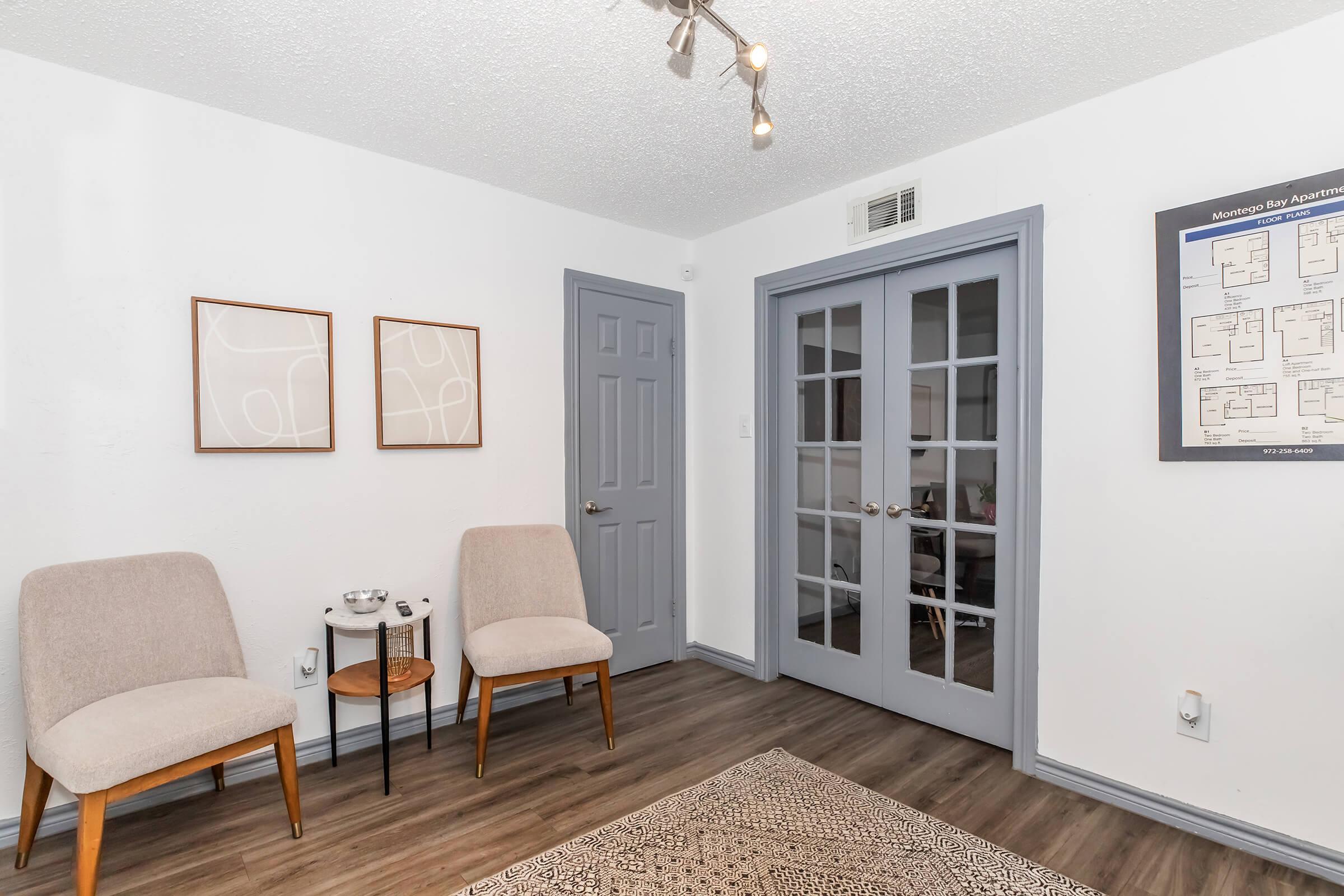 A well-decorated modern room featuring two light gray chairs with wooden legs, a small round side table, and framed abstract art on the wall. A pair of double doors with glass panes leads to another room, and a decorative rug covers the wooden floor. The walls are painted white, giving a bright and airy feel.