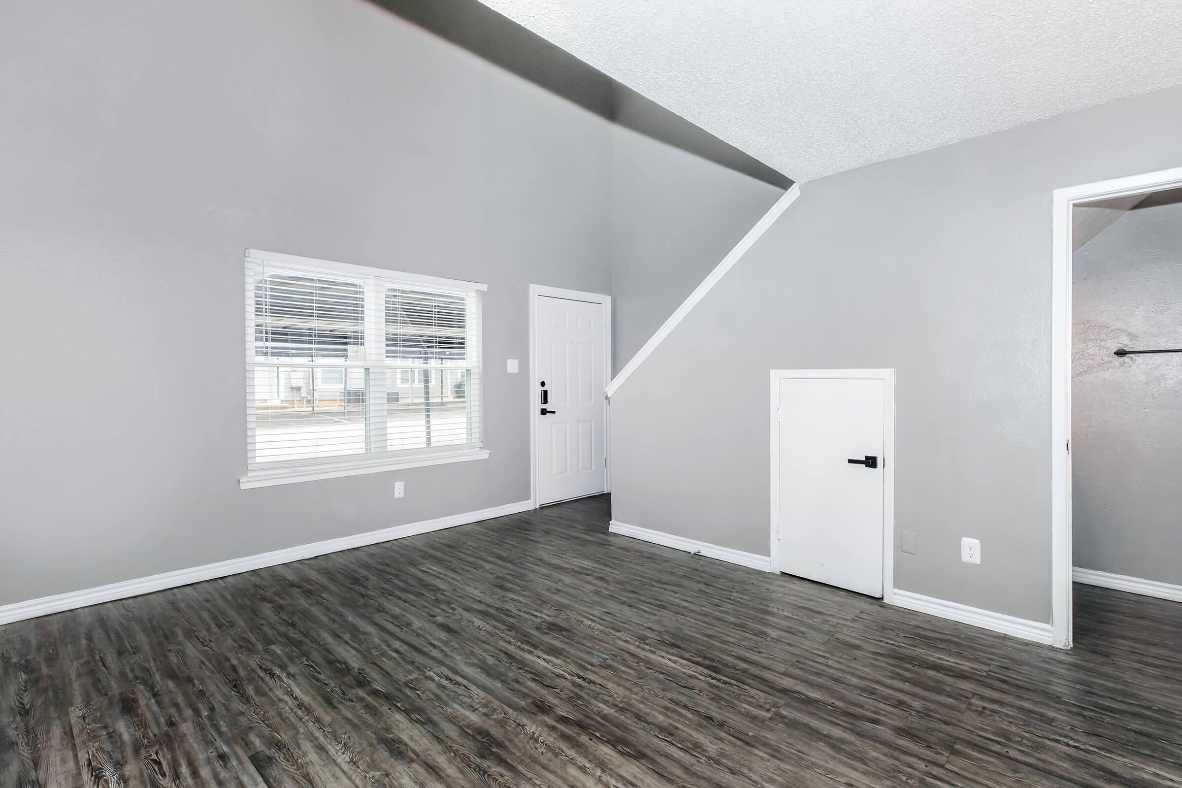 A spacious room featuring grey walls, a door leading outside, a staircase, and a small closet. Natural light filters through a window with blinds, illuminating the wooden-style flooring. The layout is open and modern, creating a welcoming atmosphere.