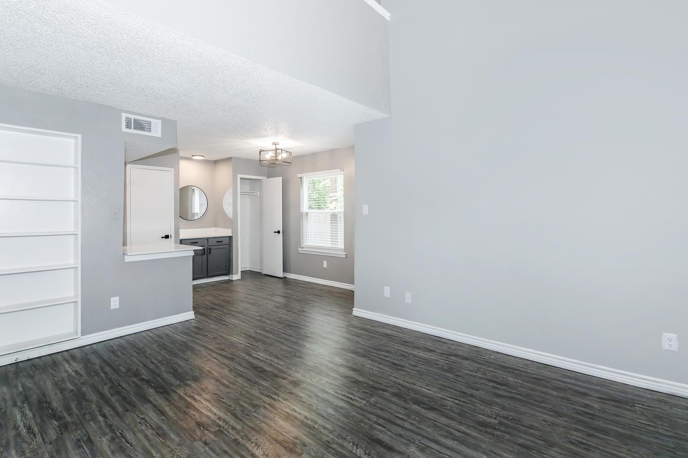 A spacious, modern living area featuring light gray walls and dark wood-like flooring. There is a built-in bookshelf on the left, a large mirror on the opposite wall, and a doorway leading to another room. Natural light streams through a window, enhancing the airy atmosphere of the room.