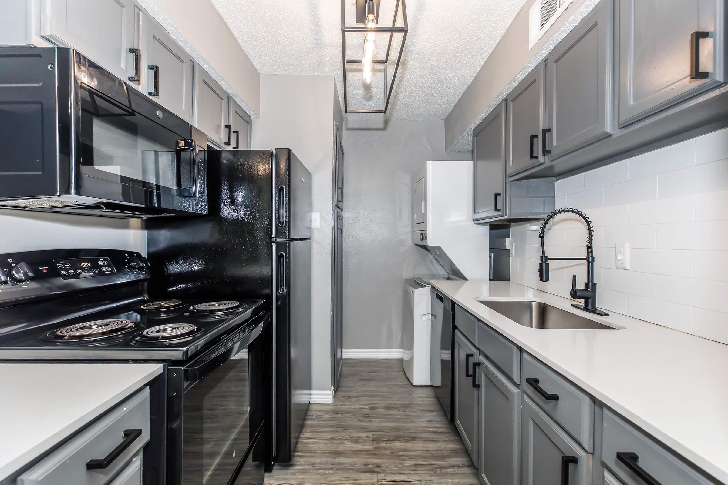 Modern kitchen featuring dark gray cabinets, stainless steel appliances, and a sleek countertop. Includes a black stove, microwave, refrigerator, and a sink with a stylish faucet. The space has a contemporary vibe with tiled walls and pendant lighting.