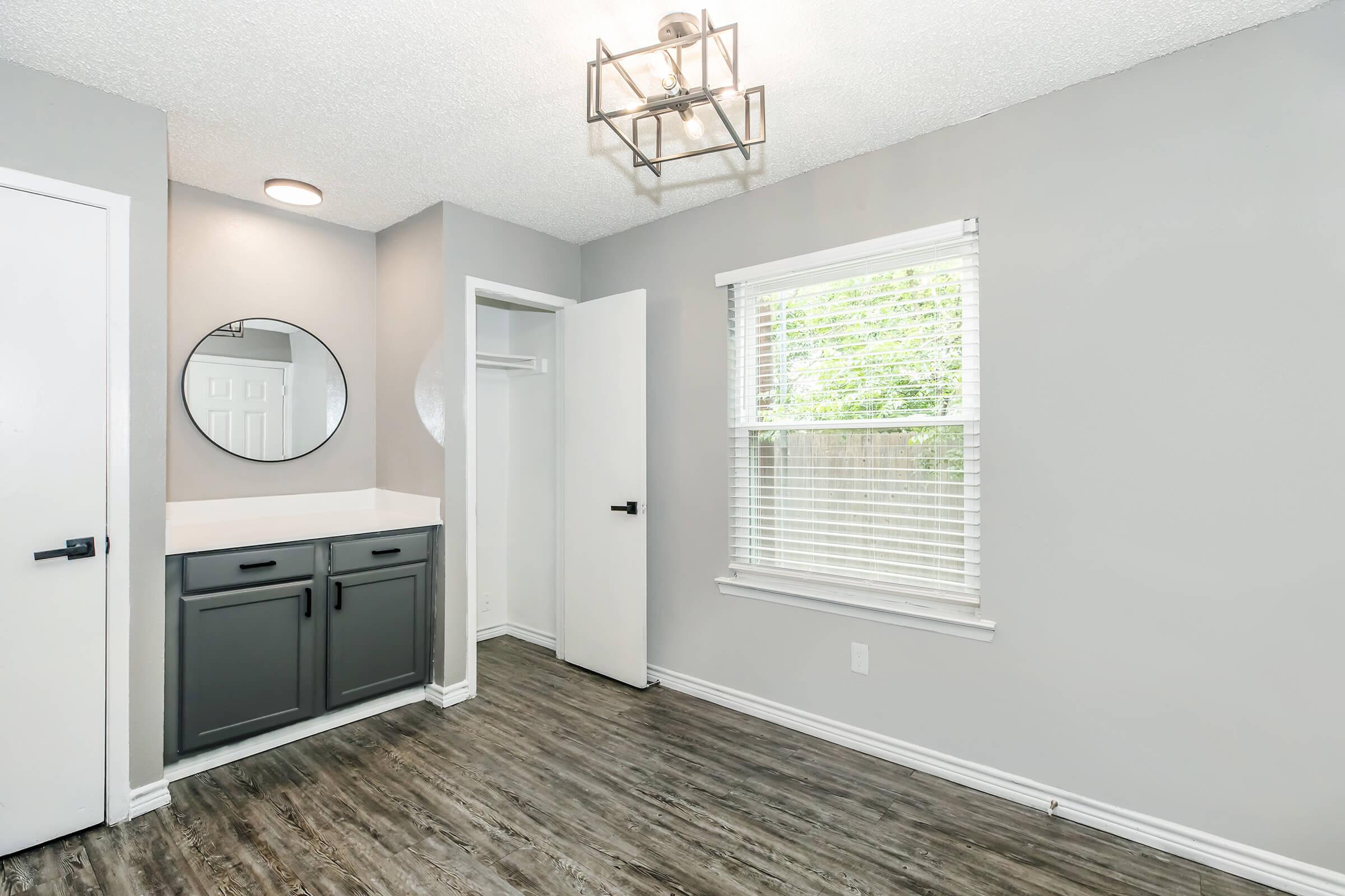 A modern, light gray room featuring a circular mirror above a vanity with dark gray cabinets, a window with white blinds allowing natural light, and an open closet door. The floor is a wood-like laminate, and a decorative light fixture hangs from the ceiling.