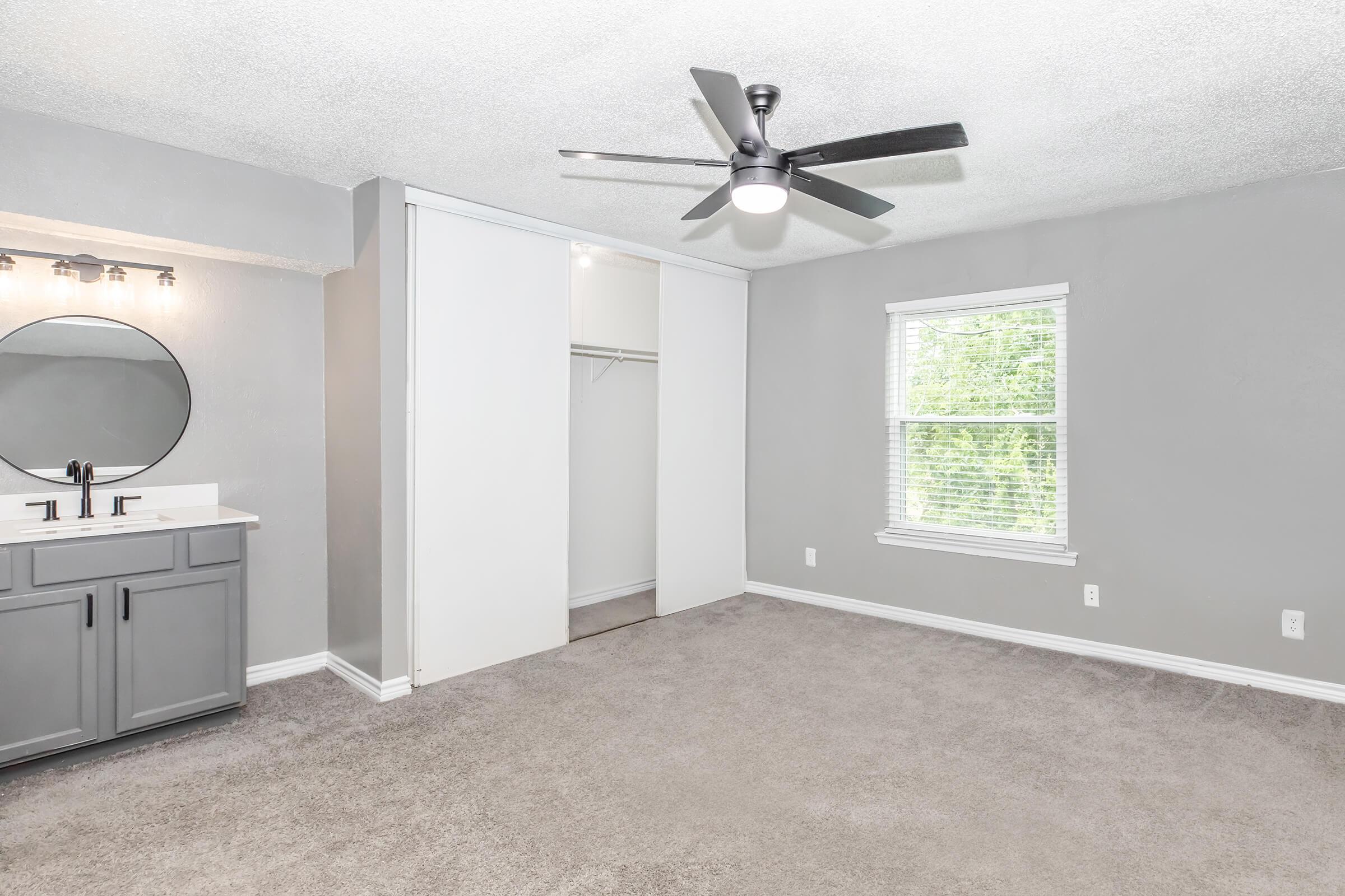 A modern bedroom featuring a gray color scheme, a ceiling fan, a large mirror above a double vanity sink, and a carpeted floor. The room has a window allowing natural light in and a sliding closet door. The walls are adorned with simple white trim, creating a clean and inviting atmosphere.