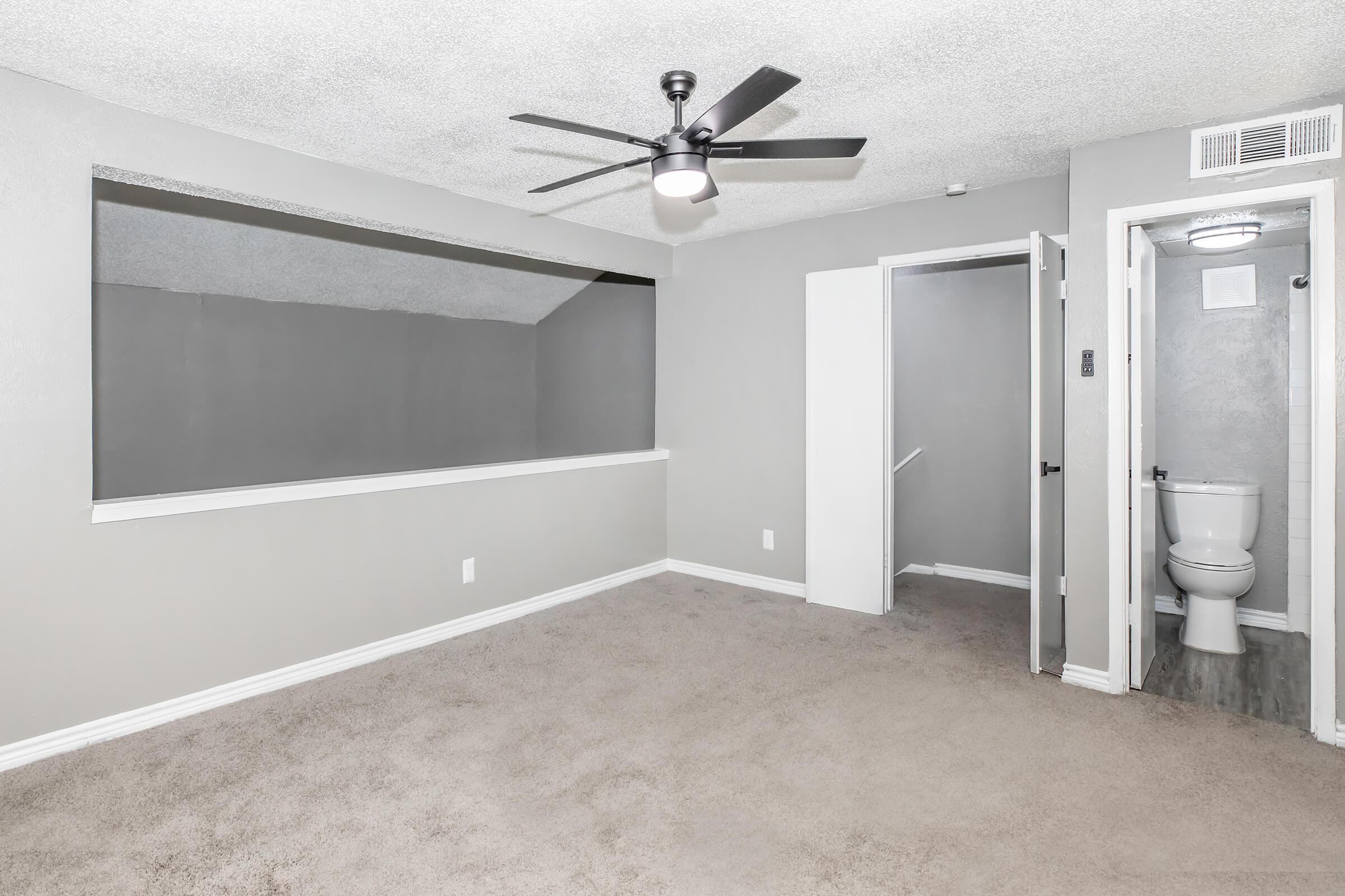 A spacious room with light gray walls and carpet, featuring a ceiling fan. There is a large window-like opening to another room, and a door leading to a bathroom area with a toilet. The layout suggests a modern apartment design, emphasizing open space and natural light.