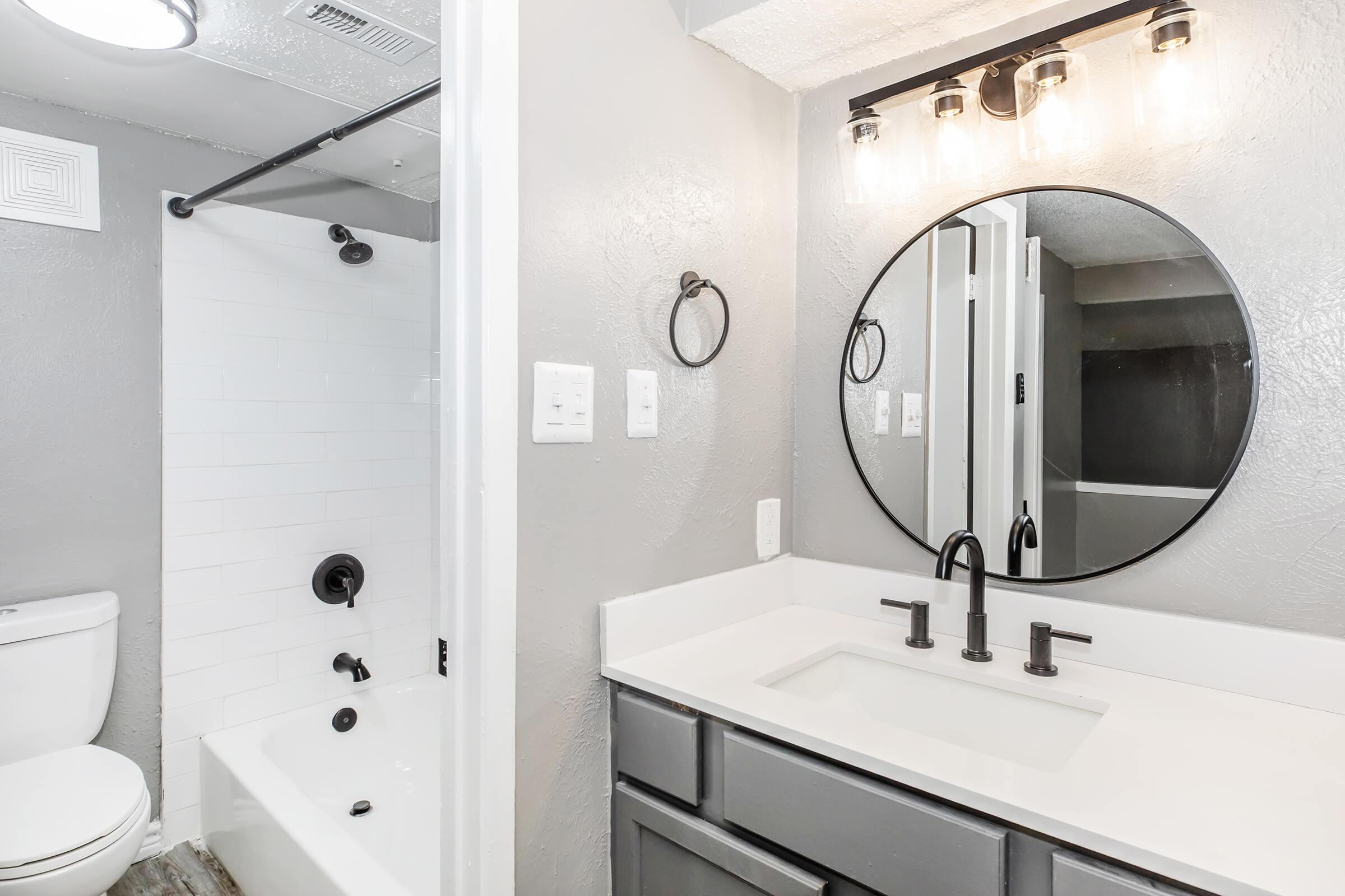 Modern bathroom featuring a circular mirror above a white countertop with black faucet fixtures. The space includes a bathtub with a shower. The walls are painted in light gray, and there are decorative light fixtures above the mirror. A toilet is visible in the corner, completing the sleek design.