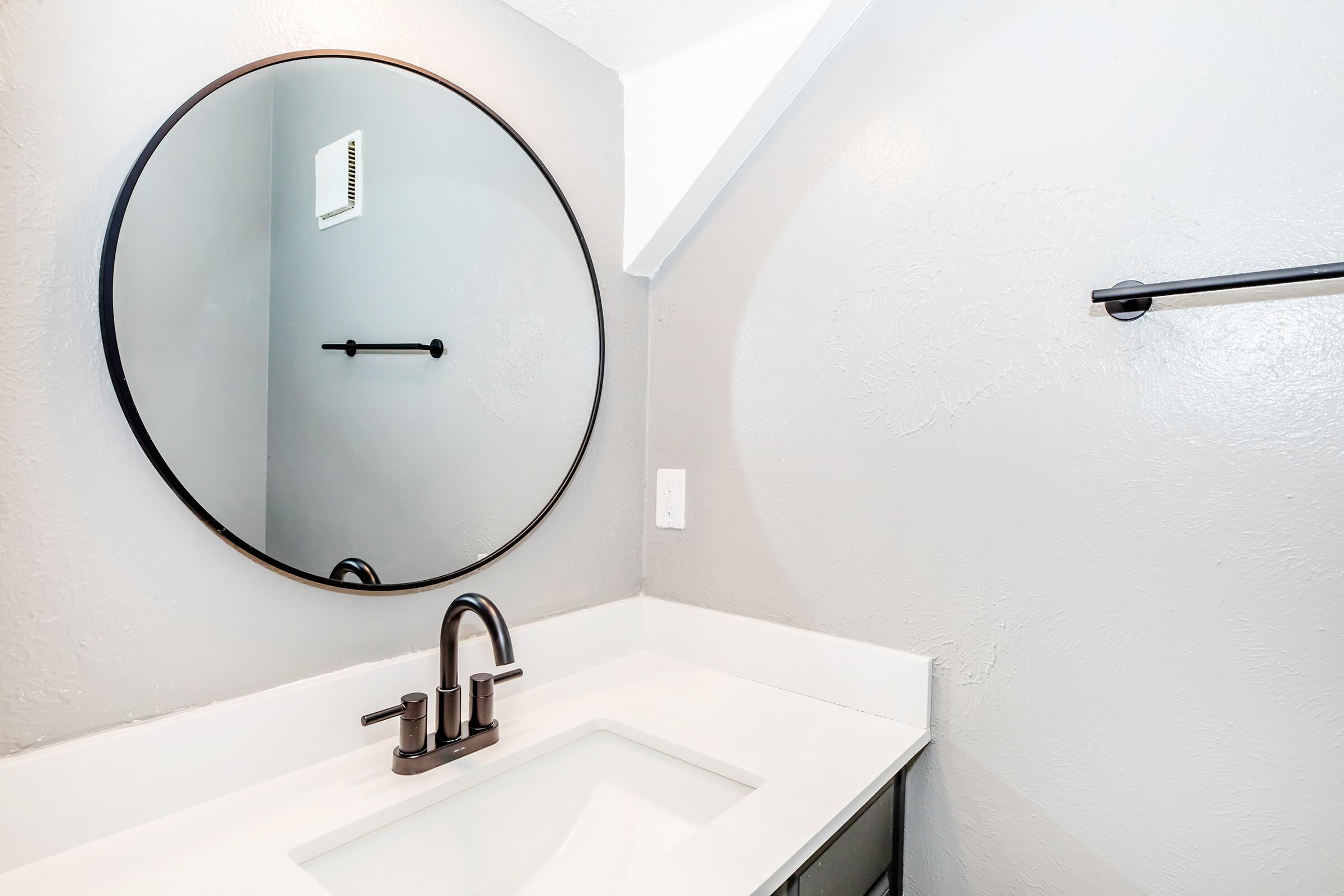 A modern bathroom with a round black-framed mirror above a white countertop sink. The wall is painted in light gray, and there's a black towel bar mounted on the wall. The overall aesthetic is clean and minimalist.