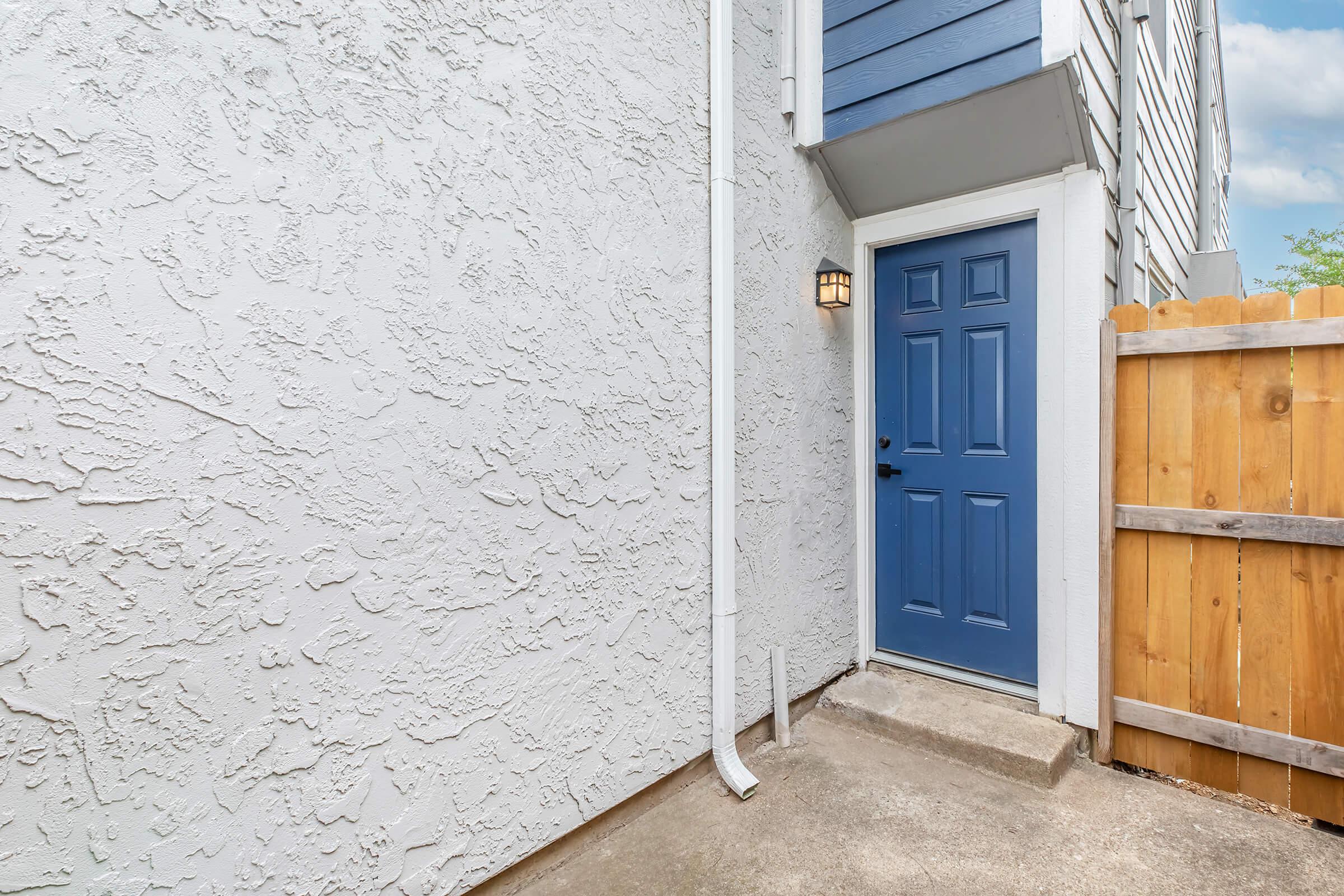 A blue front door with a lantern beside it is set against a textured white wall. The door is located next to a wooden fence, and there is a concrete path leading up to it. The overall scene is bright with a clear sky in the background.