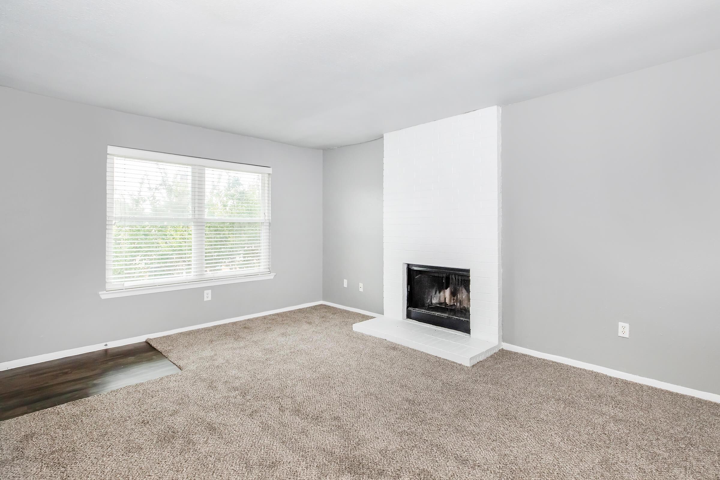 Bright living room with light gray walls, a large window letting in natural light, and beige carpet flooring. The focal point is a modern white brick fireplace on one wall, enhancing the cozy atmosphere. The space is uncluttered, offering a clean and inviting ambiance.