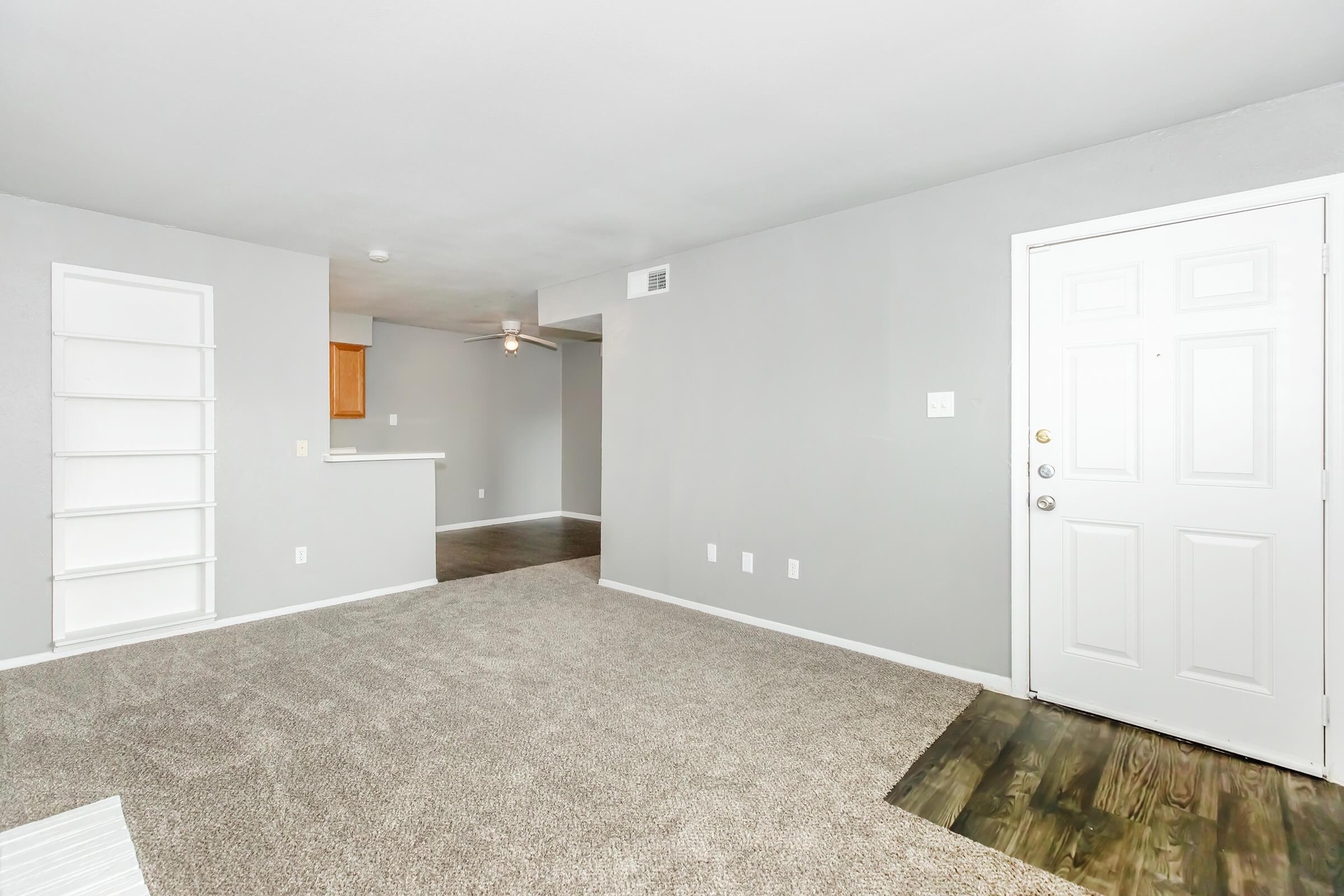 A modern, spacious living room with light gray walls and carpeted flooring. There is a front door on the right, a ceiling fan overhead, and an opening to a kitchen area in the background. A white bookshelf is mounted on the left wall, adding a touch of decor.