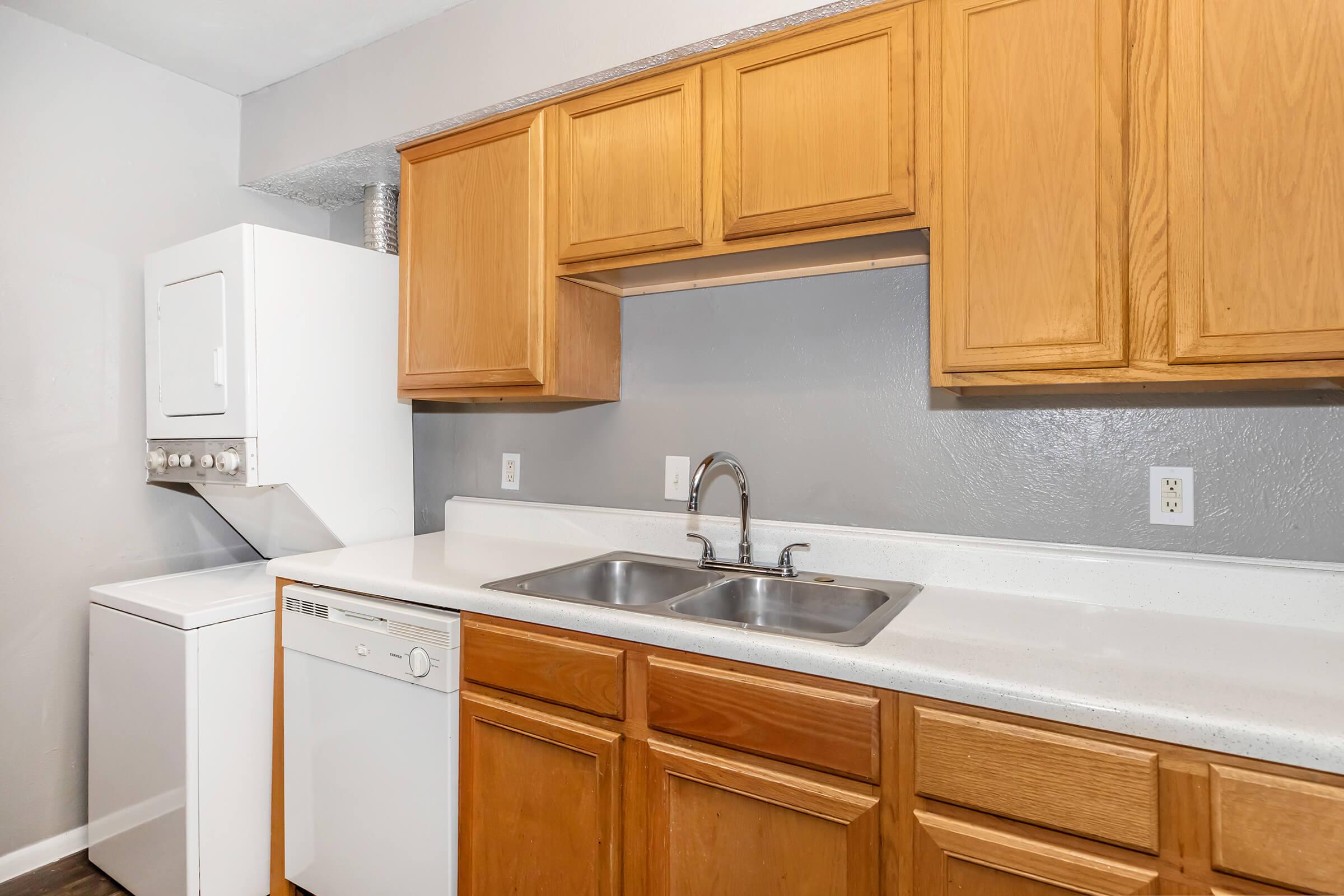 Kitchen area featuring wooden cabinetry, a stainless steel double sink, a dishwasher, and a stacked washer and dryer unit. The walls are painted gray, and the countertops have a simple white finish, creating a functional and clean look.