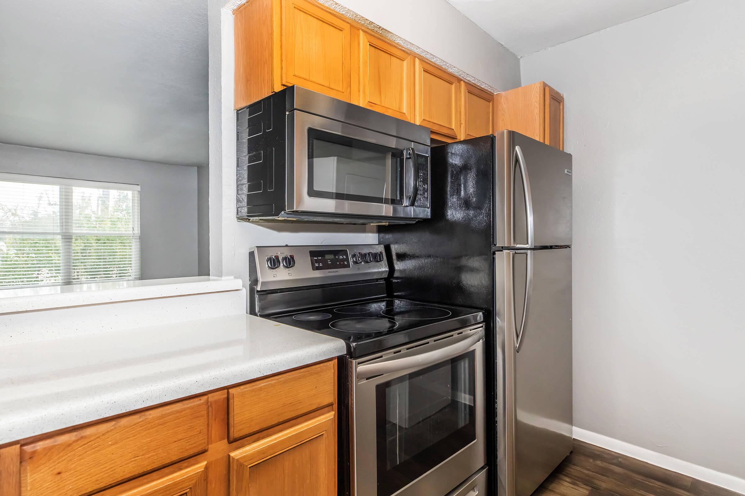 A modern kitchen featuring wooden cabinets, a stainless steel refrigerator, and a black stove with an over-the-range microwave. The countertop is light-colored, and there is a window in the background allowing natural light to enter the space.