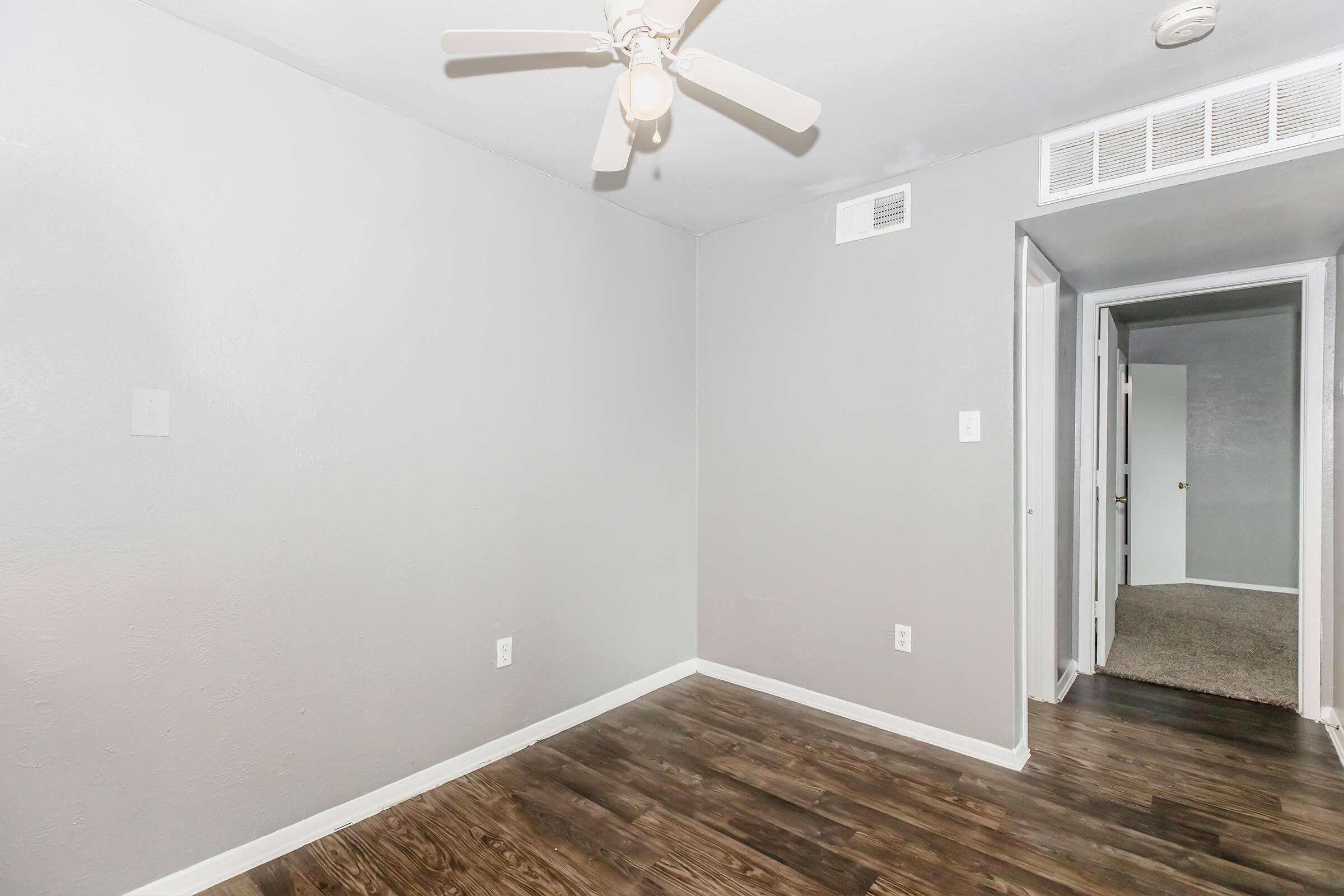 An empty room with gray walls and dark wood flooring. A ceiling fan is mounted in the center. To the right, a doorway leads to another room with beige carpet. The overall atmosphere is neutral and minimalistic.