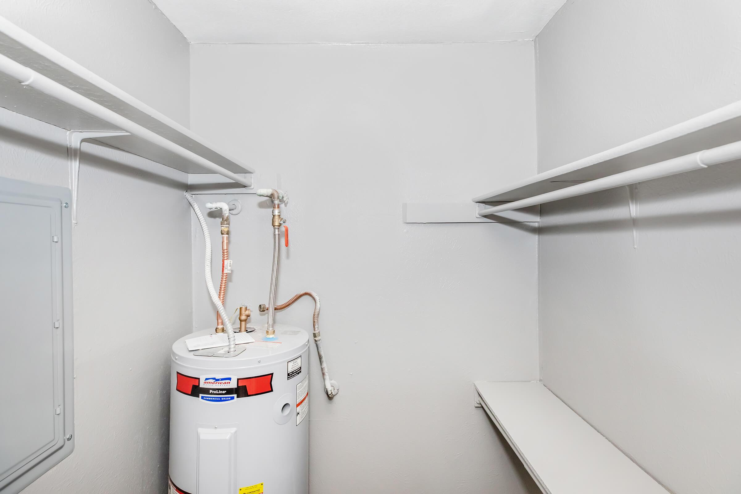 A small utility closet with gray walls, featuring a water heater on the floor. The water heater has various pipes and connectors attached. Two shelves are installed on opposing walls, one of which is empty and the other is partially filled with pipe fixtures. The space appears clean and organized.