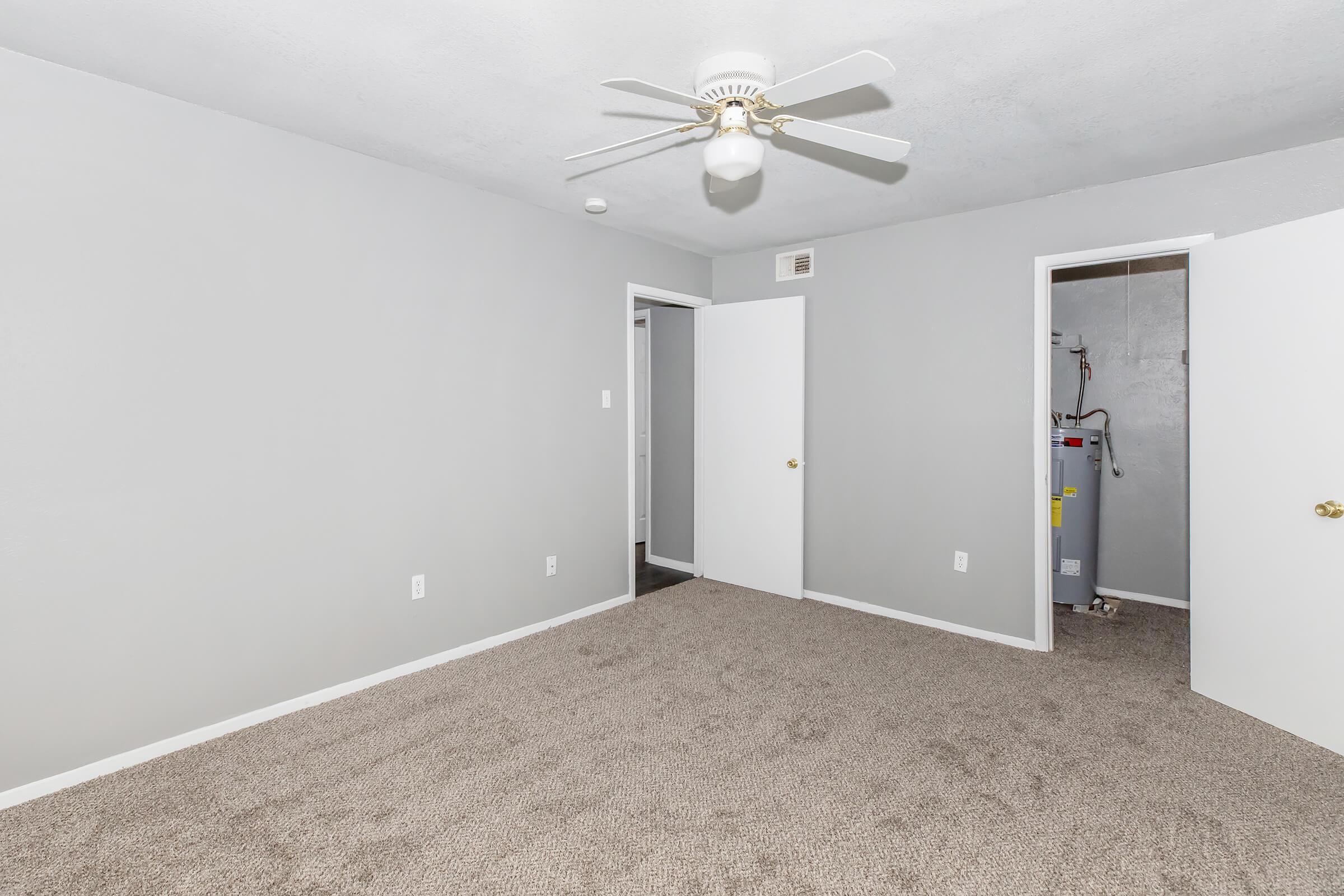 A light gray room with freshly painted walls and beige carpet. A ceiling fan is mounted in the center, and there are two white doors on the right leading to other spaces. One door is slightly ajar, revealing a glimpse of a utility area. The room is well-lit and appears clean and empty, suitable for various uses.